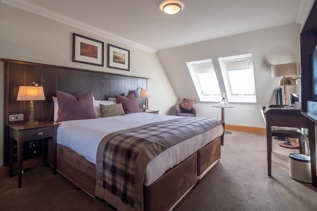A modern hotel room with a large bed, two bedside tables with lamps, a desk with a chair, and two windows providing natural light.