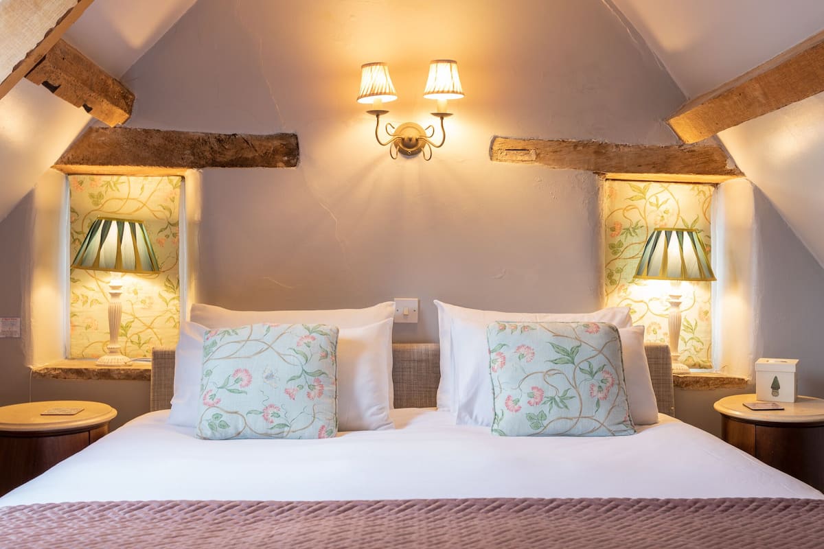 A bedroom with a sloped ceiling, two wall-mounted lamps, a bed with floral-patterned pillows, and a nightstand with a lamp.