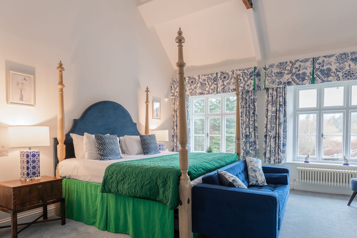 A four-poster bed with a green bedspread and blue headboard, a blue armchair, a wooden nightstand, and a window with floral curtains.