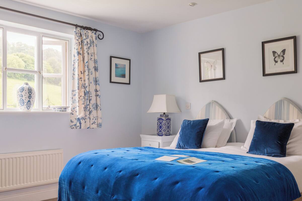 A bedroom with a large bed, two bedside tables, a window with curtains, and two framed pictures on the wall.