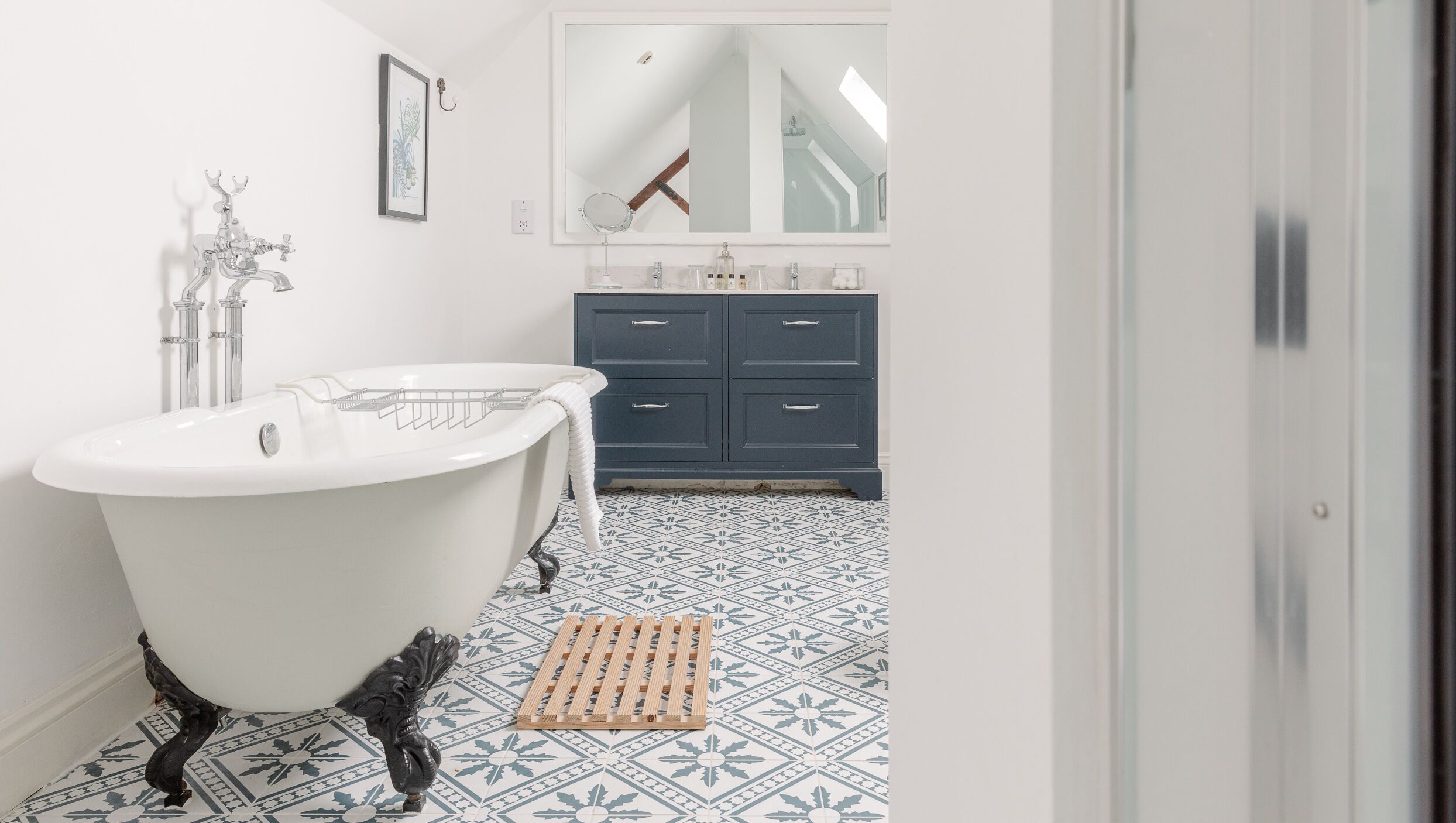 A bathroom with a clawfoot bathtub, a wooden bench, a blue cabinet with drawers, and a patterned floor.
