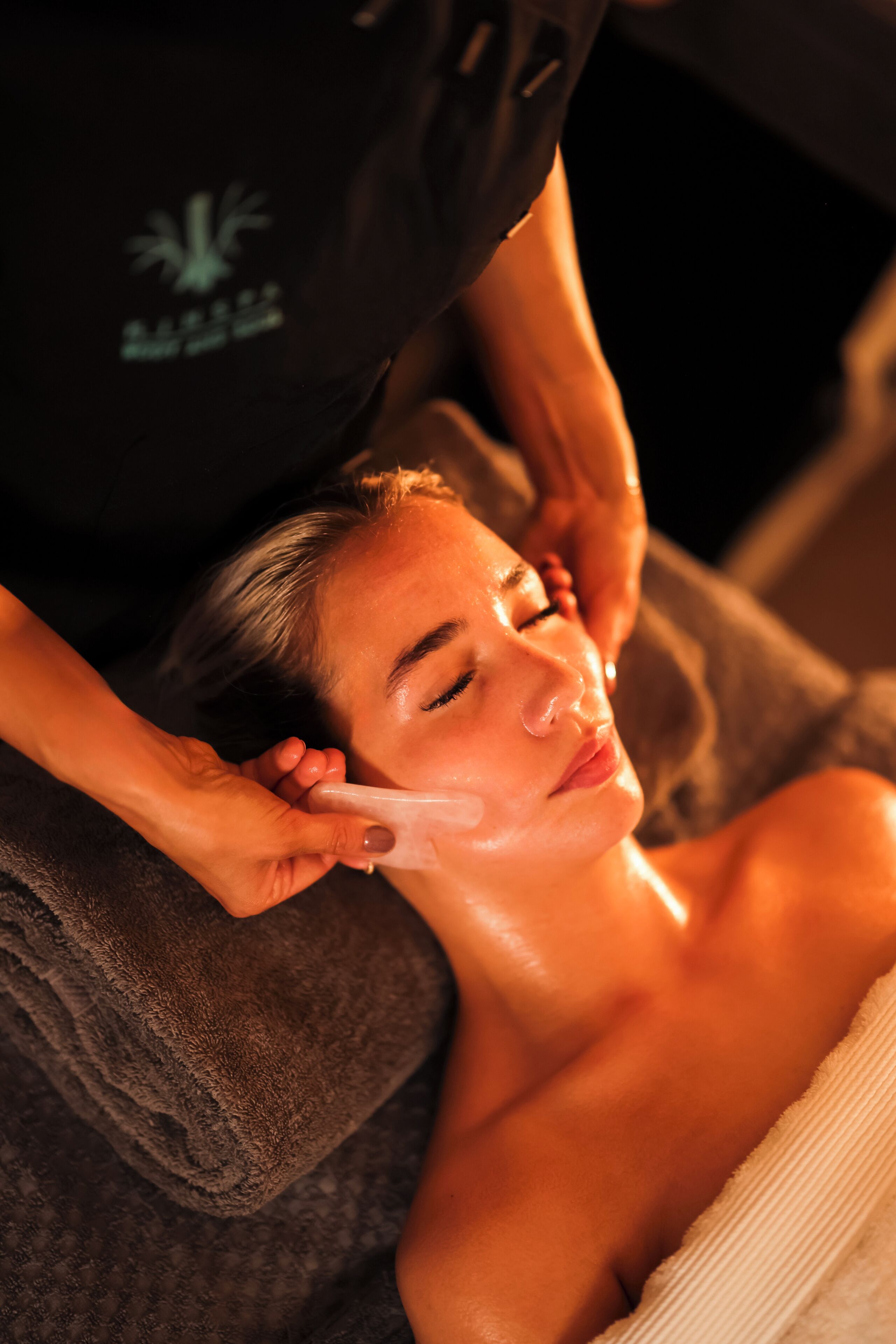 A person receiving a facial treatment in a room with a dark-colored towel and a logo on the wall.