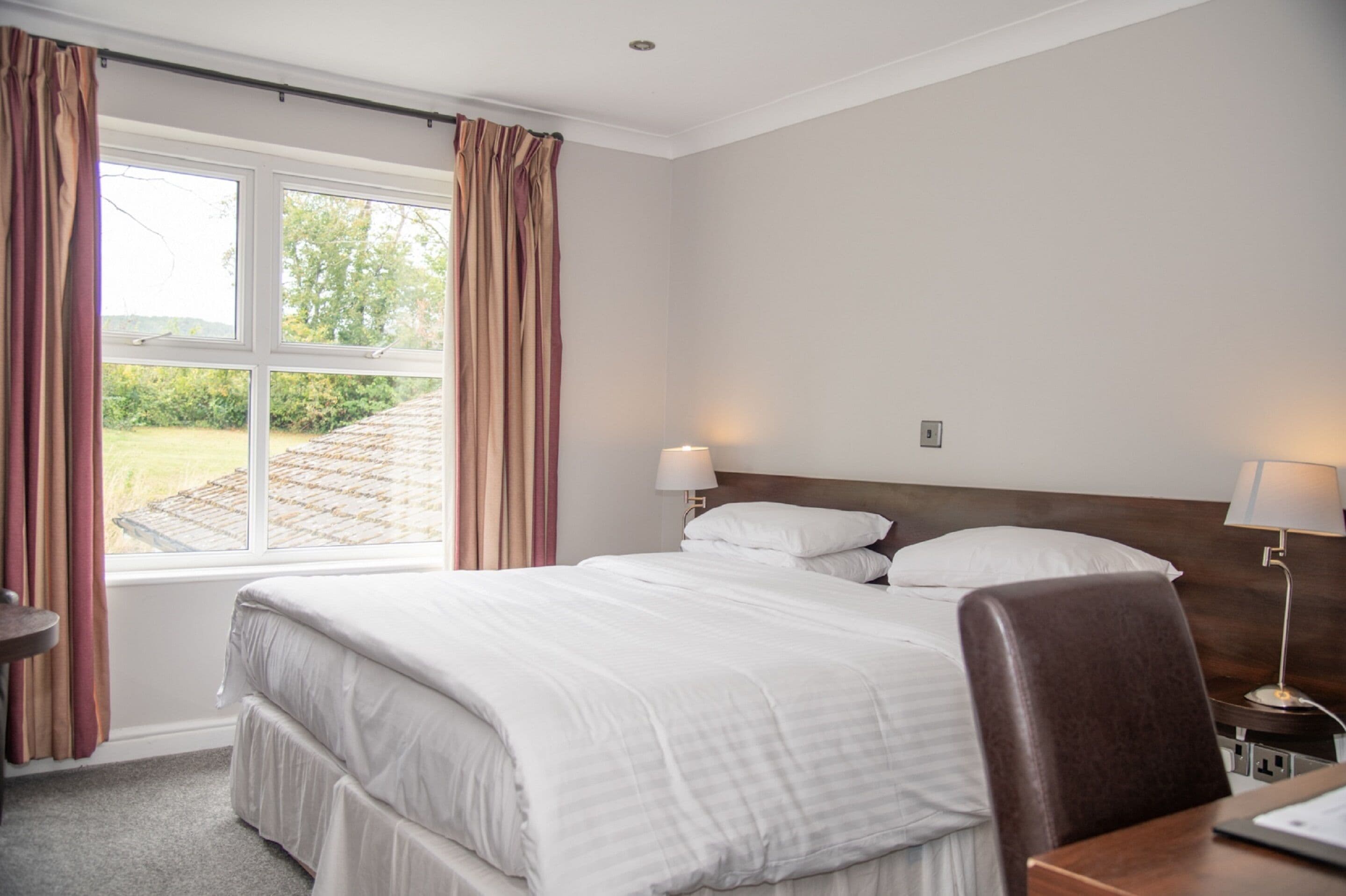 A hotel room with a large bed, two bedside lamps, a wooden headboard, a desk with a chair, and a window with curtains.