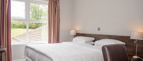 A hotel room with a large bed, two bedside lamps, a wooden headboard, a desk with a chair, and a window with curtains.