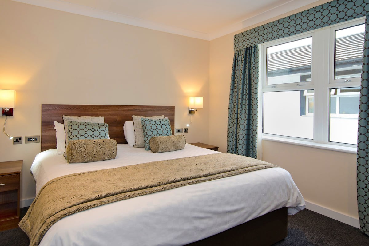 A hotel room with a large bed, two bedside lamps, a window with curtains, and a headboard with a geometric pattern.