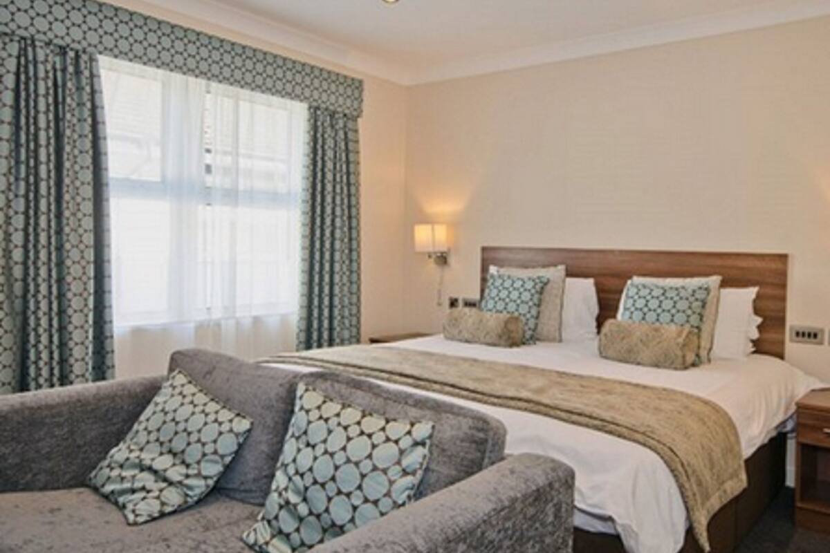 A hotel room with a bed, a nightstand, a lamp, a sofa with patterned cushions, and a window with patterned curtains.