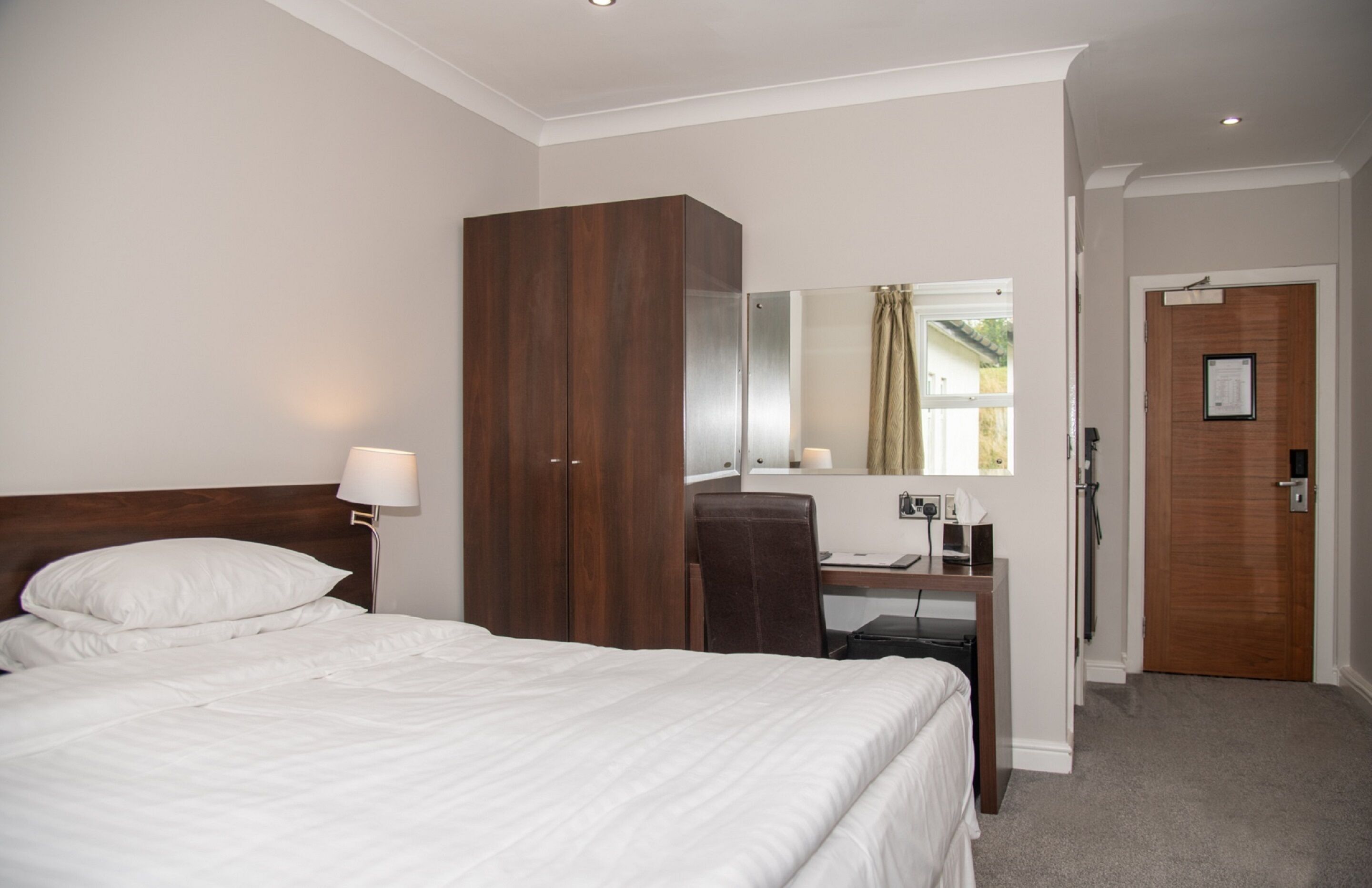 Ein Hotelzimmer mit Bett, Schreibtisch, Stuhl, Kleiderschrank, Spiegel und Tür.