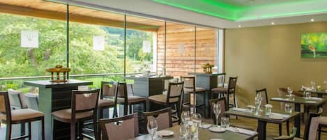 A restaurant with tables set for dining, a bar area, and large windows offering a view of greenery.