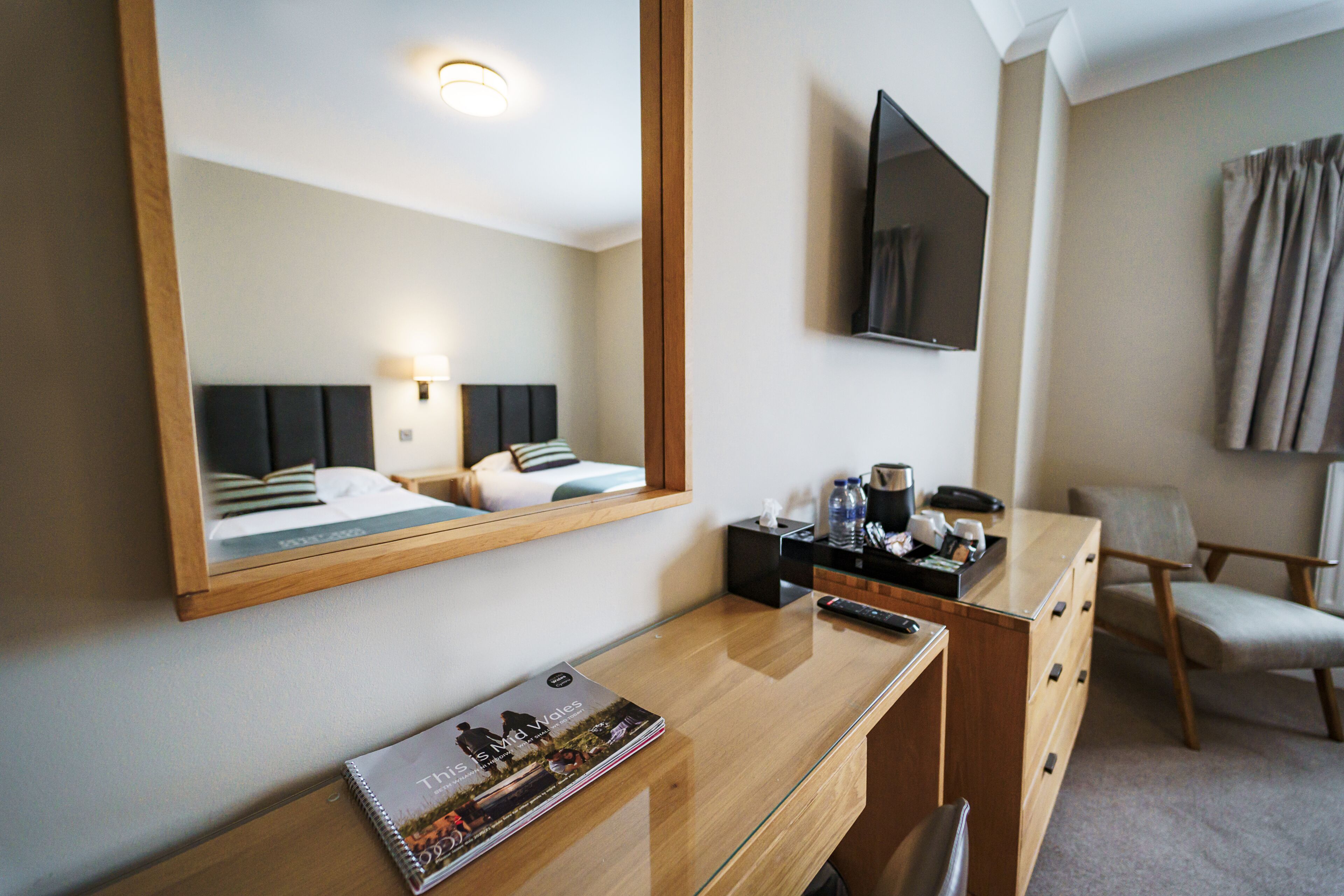 A hotel room with a wooden desk, a mirror, a television, a bed with a headboard, a chair, and a magazine on the desk.