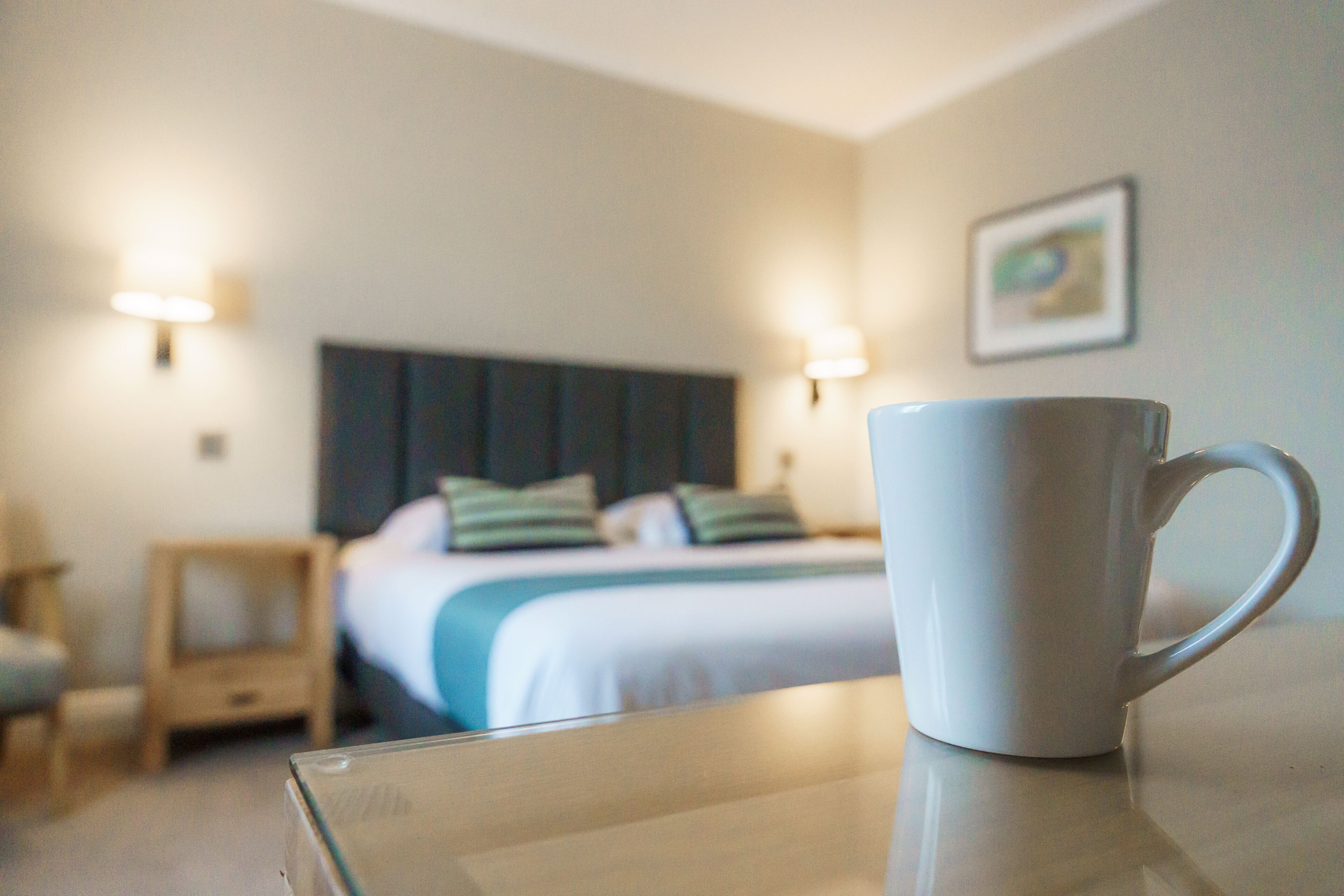 A hotel room with a bed, bedside tables, a white mug on a table, and wall-mounted lights.