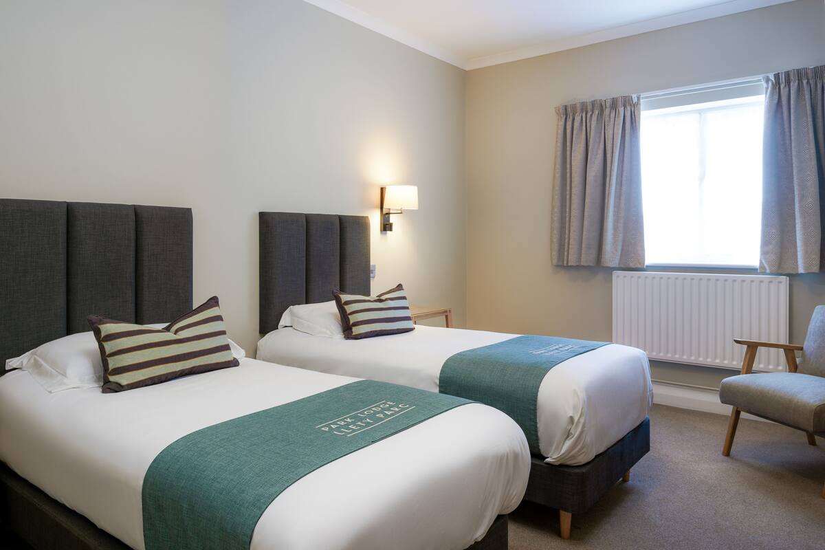 A hotel room with two beds, a nightstand, a lamp, a chair, and a window with curtains.