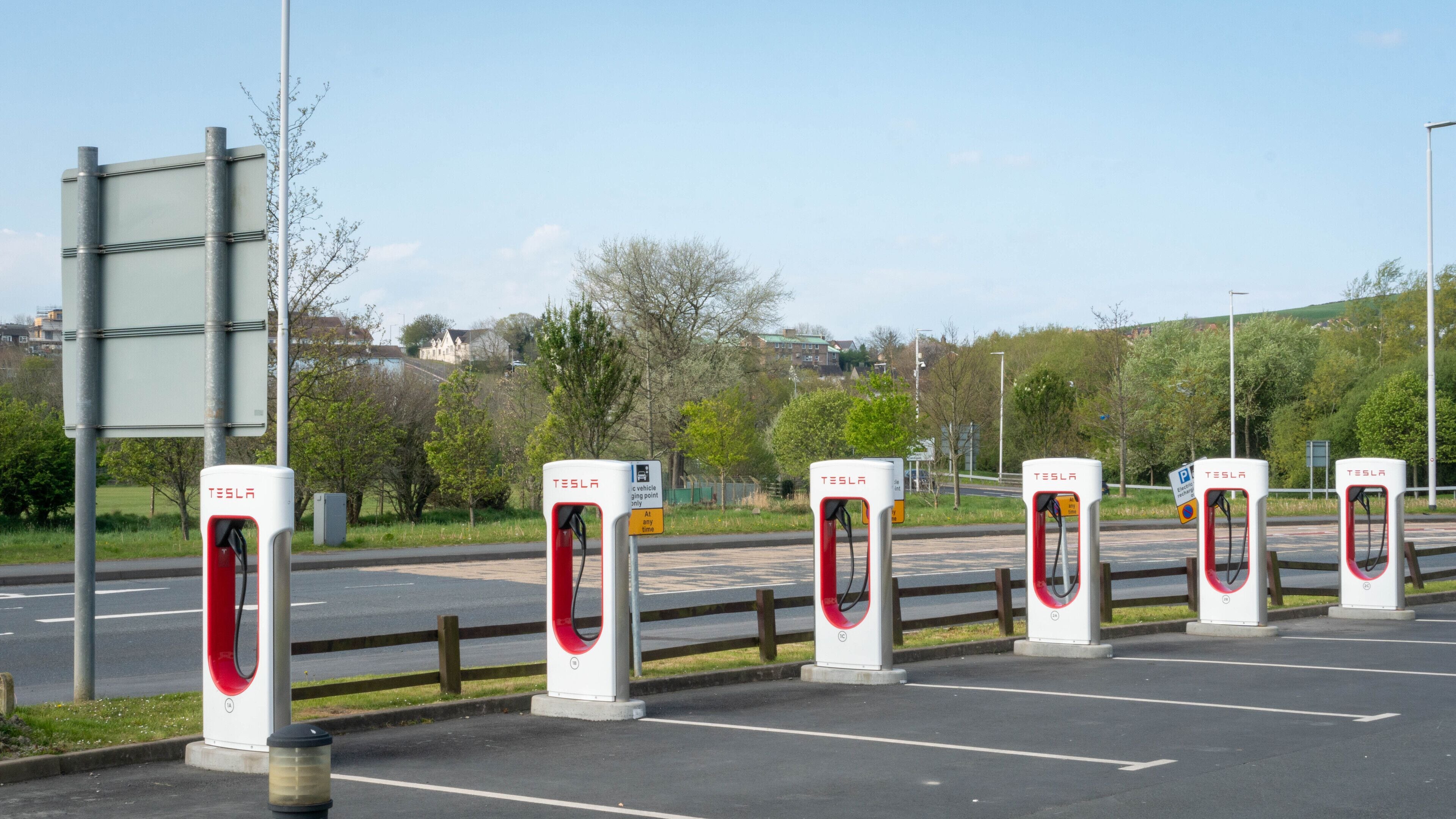 A row of Tesla charging stations along a road with a fence and greenery in the background.