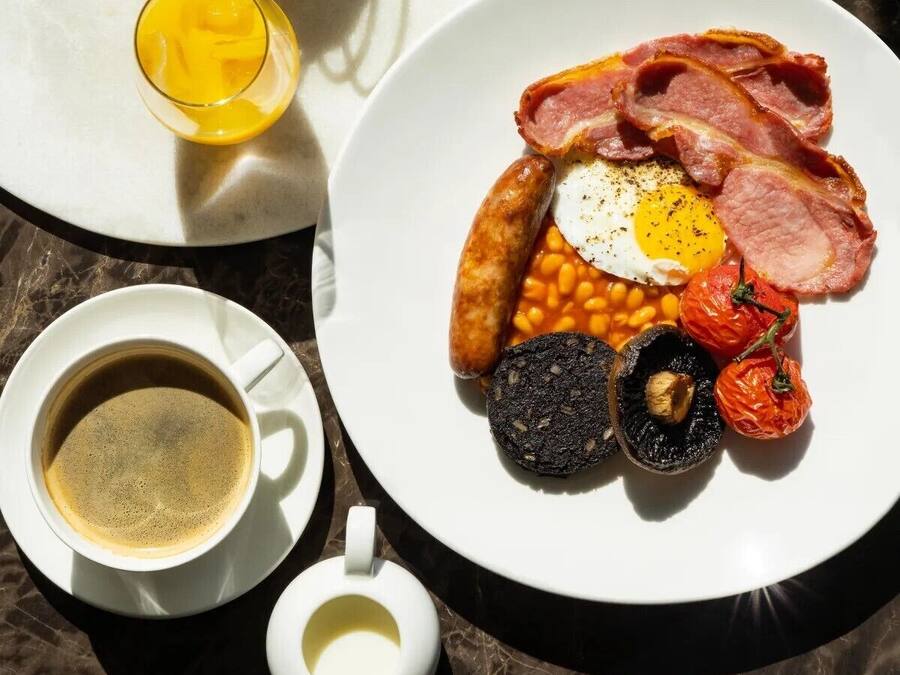 Een ontbijt bord met spek, eieren, bonen, tomaten en bloedworst, vergezeld van een kop koffie en een glas jus d'orange.