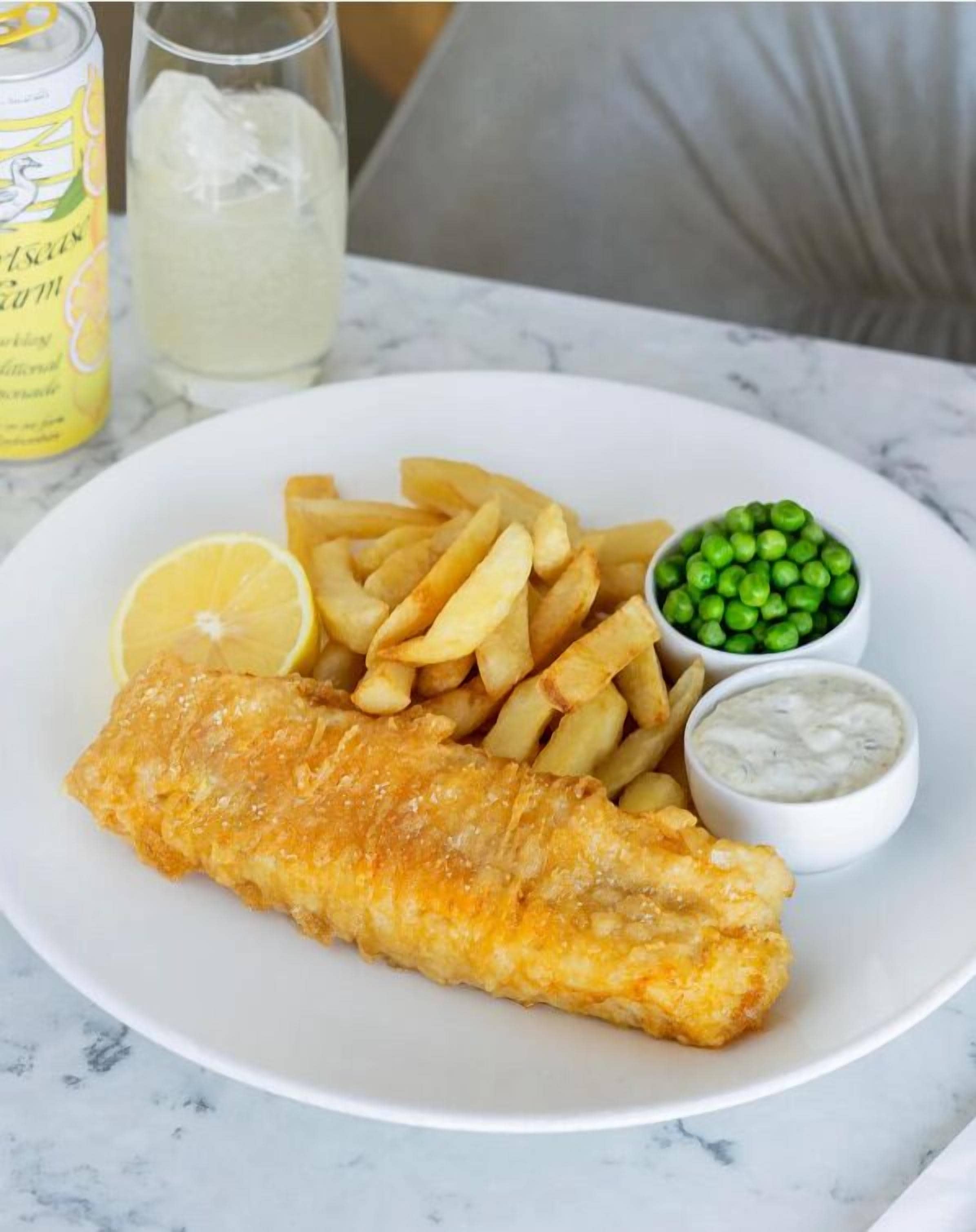 A plate with fish and chips, lemon, peas, and two dipping sauces.