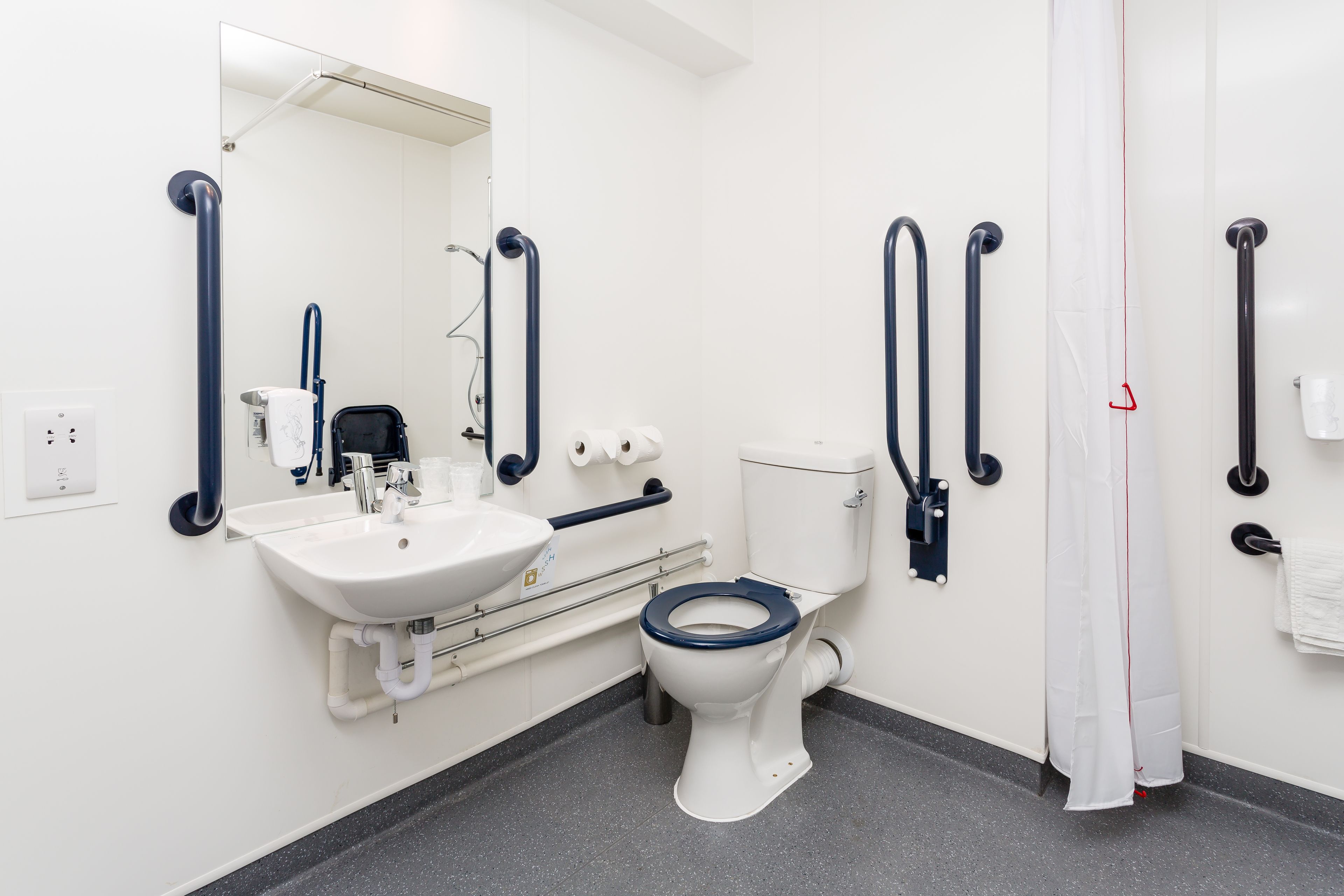 Ein modernes Badezimmer mit Waschbecken, Toilette und Dusche in Weiß. Es sind Haltegriffe und ein Spiegel vorhanden.