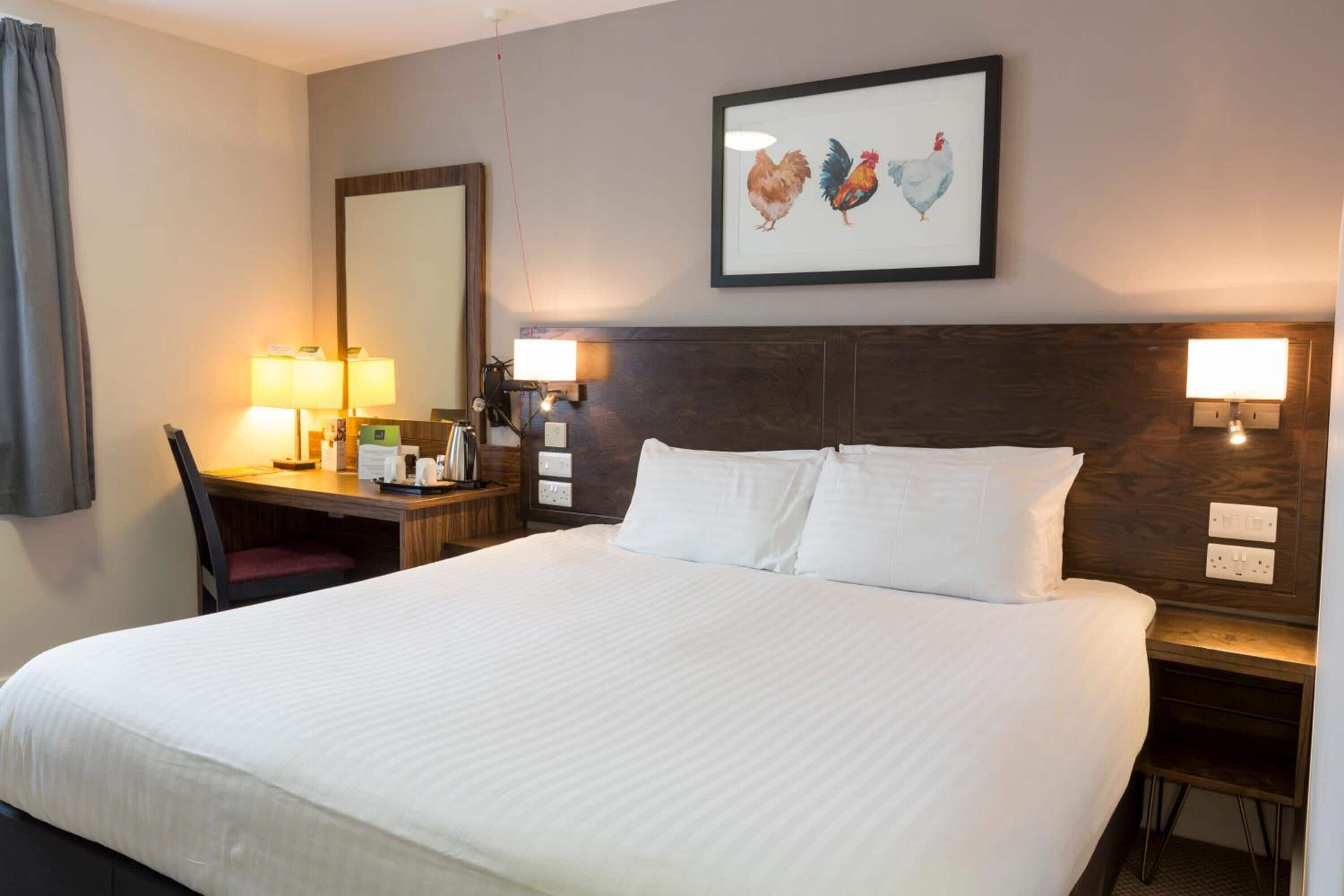 Habitación de hotel con una cama grande, un escritorio con silla, dos lámparas de noche y un cuadro en la pared.