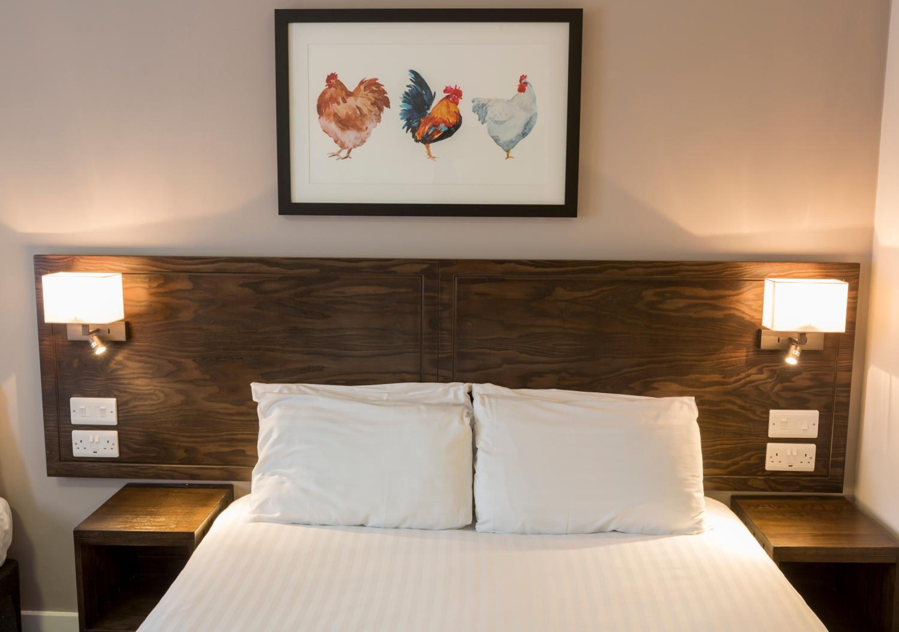 Una habitación de hotel con una cama grande, dos mesitas de noche y una obra de arte enmarcada con gallinas en la pared.