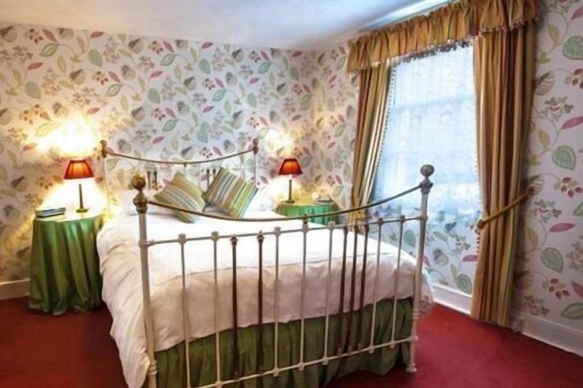 A bedroom with a four-poster bed, floral wallpaper, two bedside lamps, and a window with curtains.