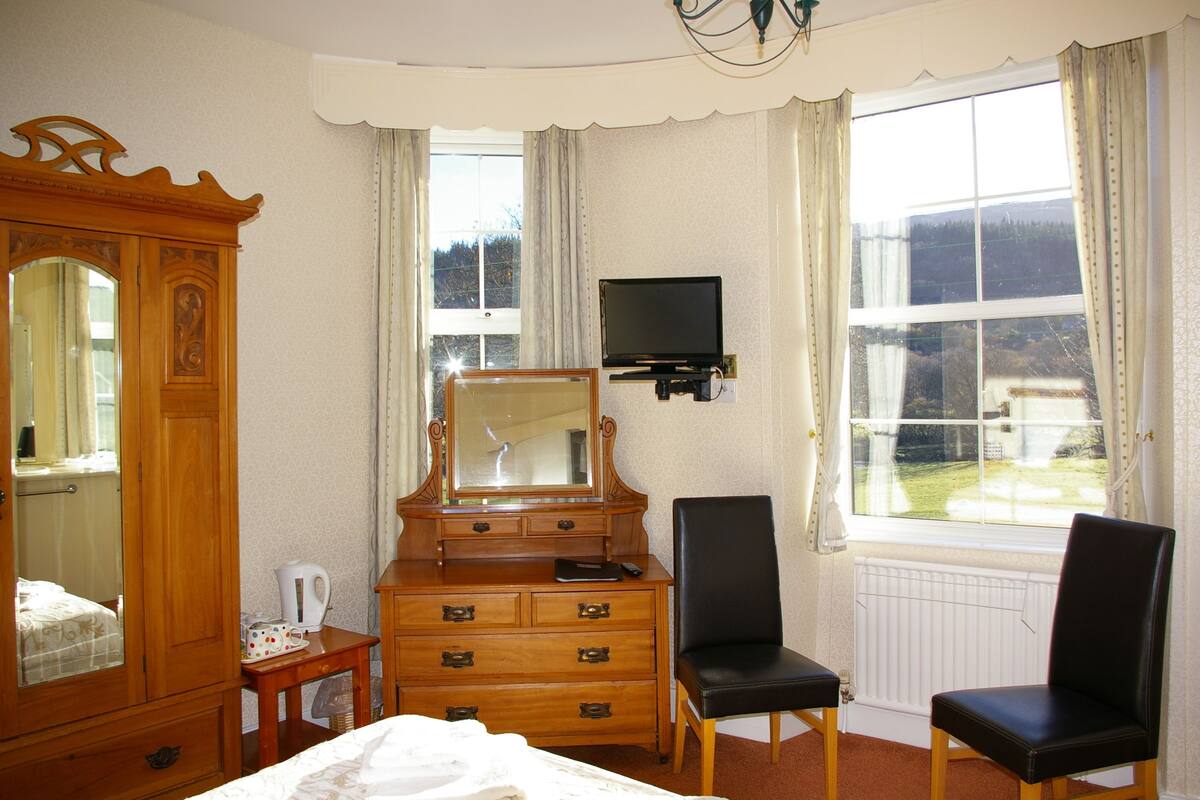 Habitación de hotel con cómoda de madera, un espejo, televisor montado en la pared, dos sillas negras y un ventanal con cortinas.