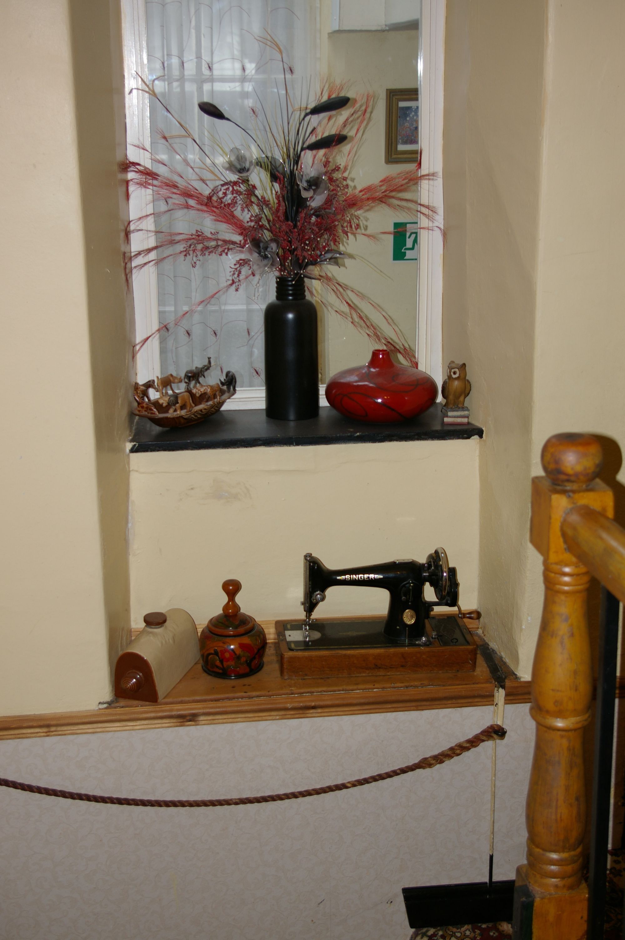 Una máquina de coser antigua sobre una mesa de madera, un florero con plantas secas y un pequeño búho de madera en un estante.
