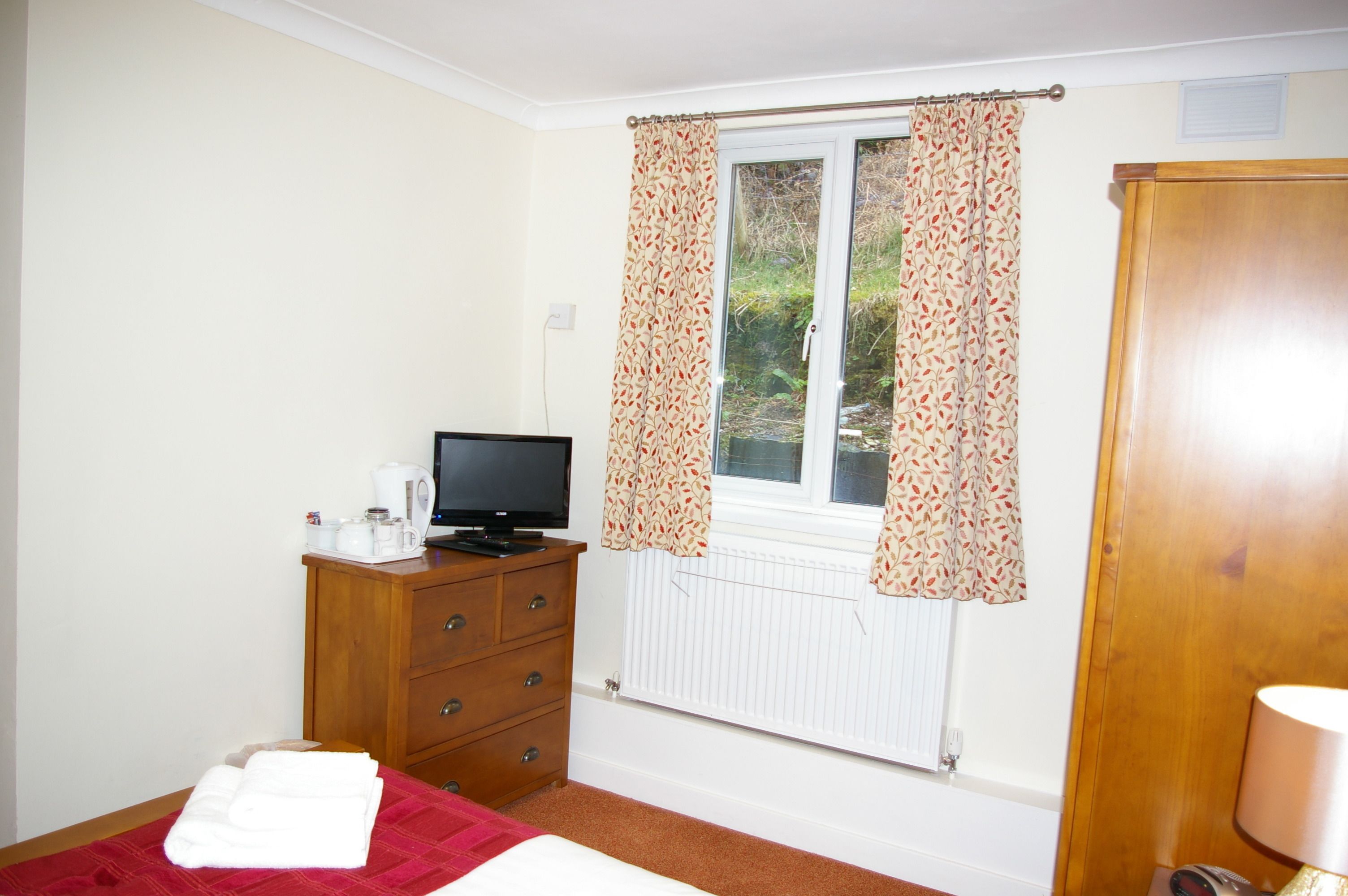 Une chambre à coucher avec un lit, un téléviseur posé sur une commode en bois, un radiateur et une fenêtre avec des rideaux.