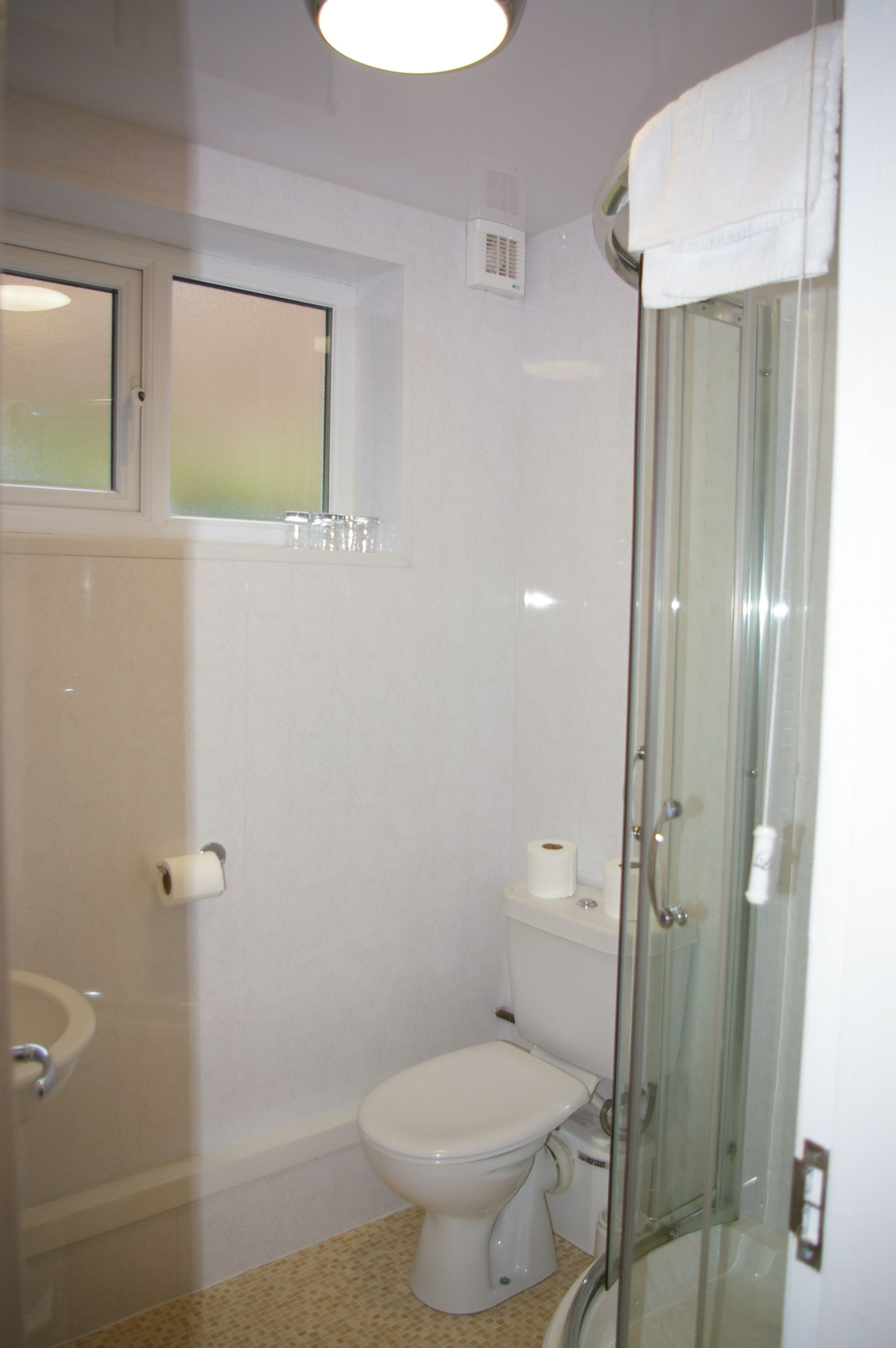 Une petite salle de bain blanche, avec des toilettes, une douche à parois de verre et une fenêtre située au-dessus des toilettes.