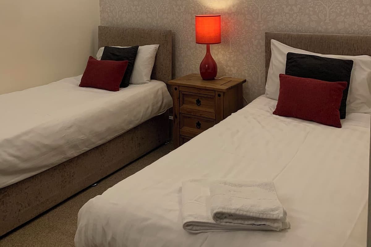 Habitación de hotel con dos camas individuales, cada una con ropa de cama blanca y almohadas rojas. Hay una mesita de noche de madera con una lámpara roja entre las camas. Las paredes tienen un estampado de papel tapiz floral, y hay dos luminarias fijadas a la pared.