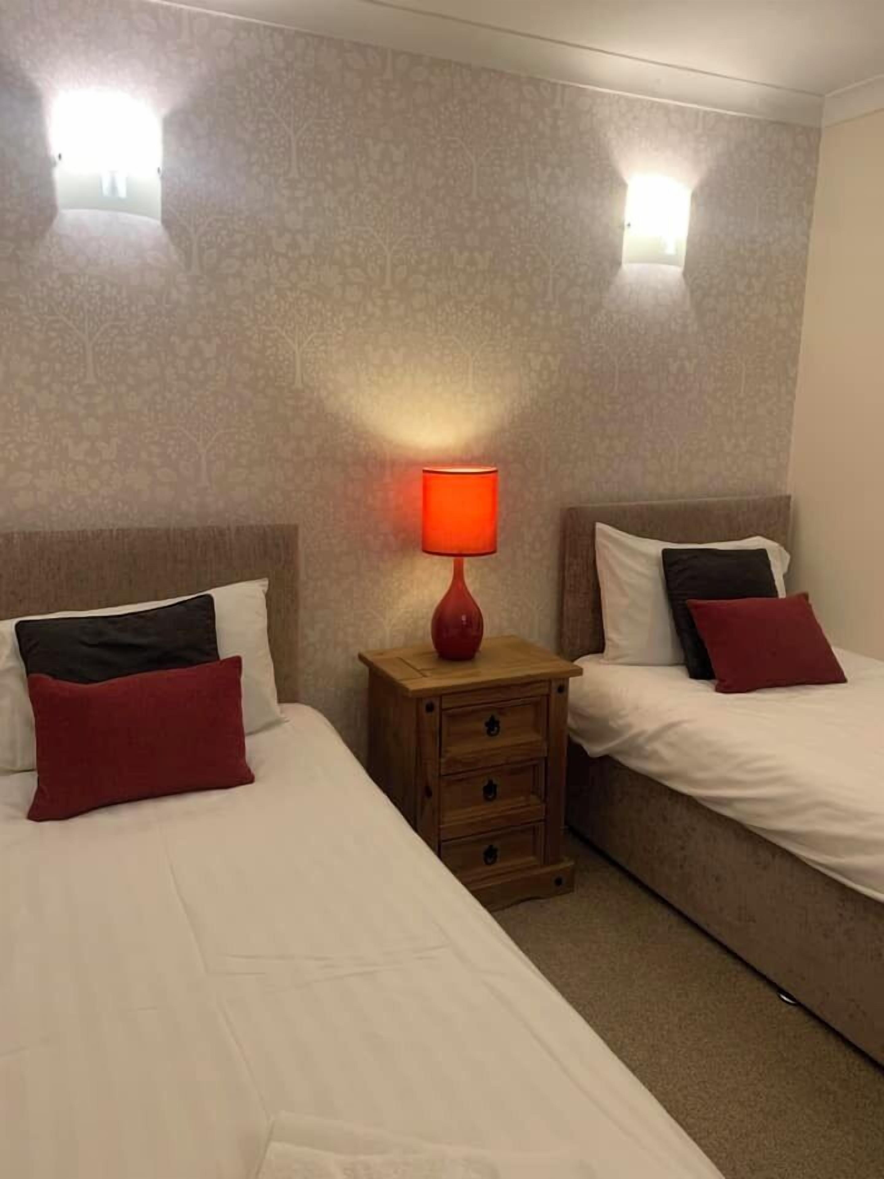 Une chambre d’hôtel avec deux lits, chacun agrémenté de draps blancs et d’oreillers rouges. Une table de chevet en bois avec une lampe rouge est placée entre les deux lits. Les murs sont recouverts d’un papier peint texturé, et deux lampes murales y sont fixées.