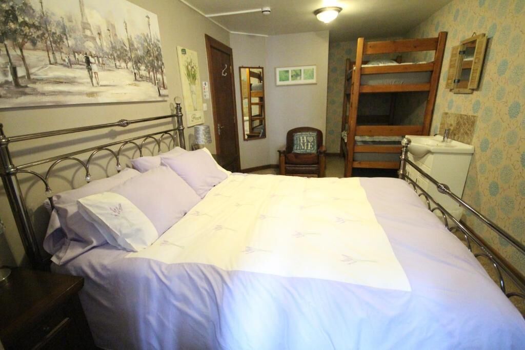 A bedroom with a metal bed frame, a bed with a white and lavender bedspread, a wooden chair, a wooden bunk bed, and a wall-mounted mirror.