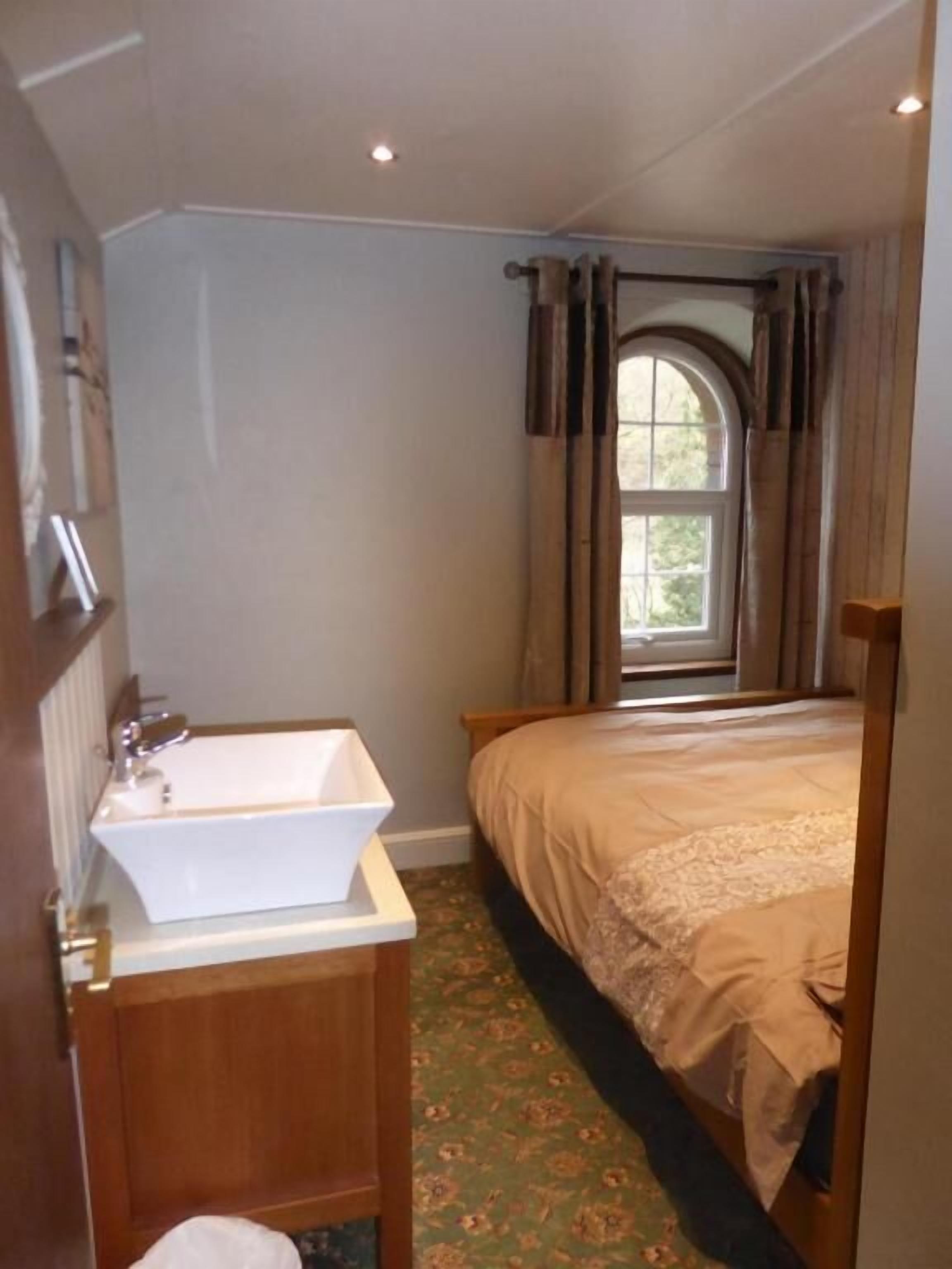 A bedroom with a single bed, a window with curtains, a bedside table, and a bathroom with a sink.