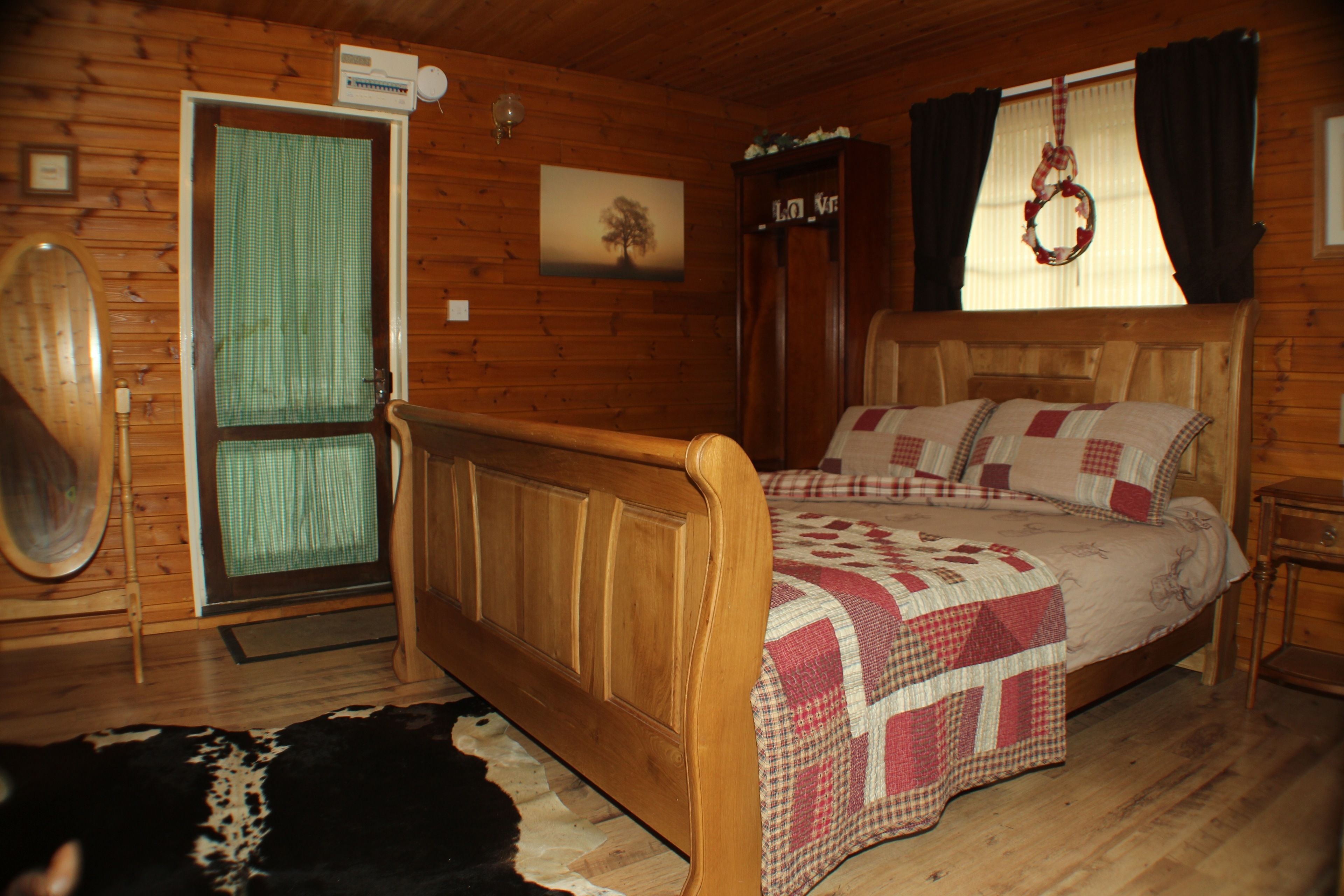 A wooden cabin bedroom with a bed, a wooden headboard, a door with a green curtain, a wall-mounted air conditioner, and a window with a wreath.