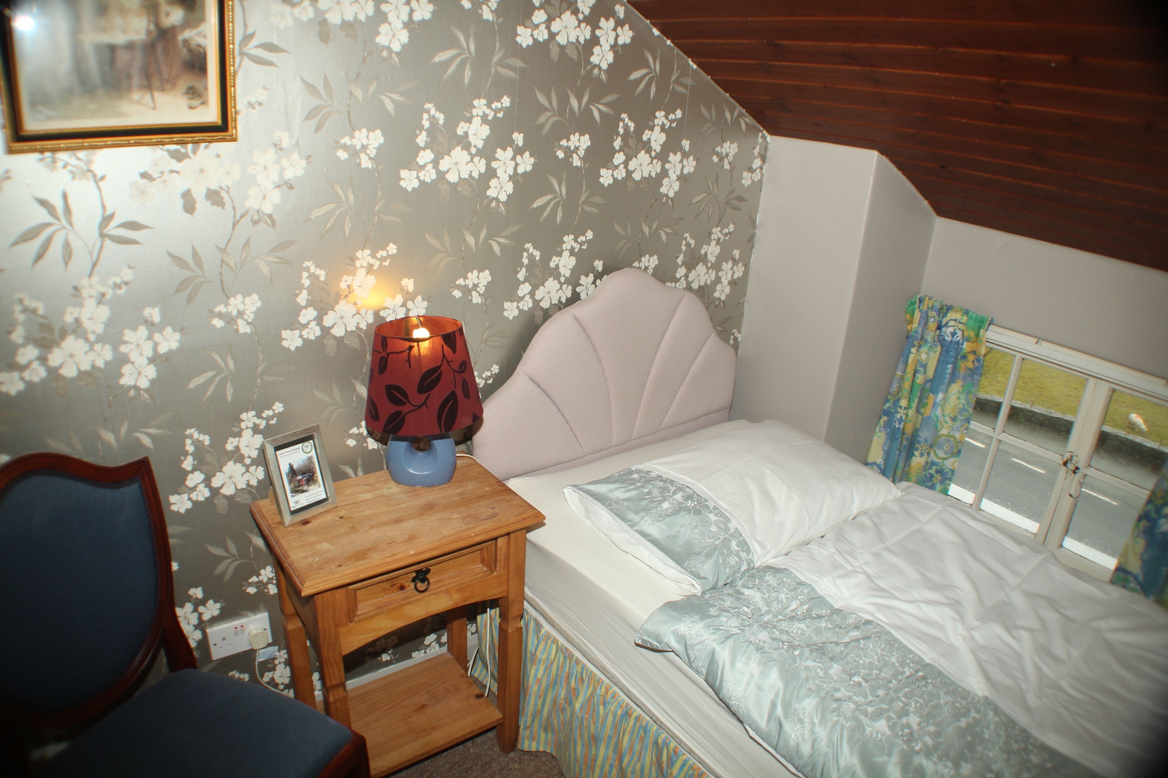 A bedroom with a bed, a bedside table with a lamp, a chair, and a window with curtains.