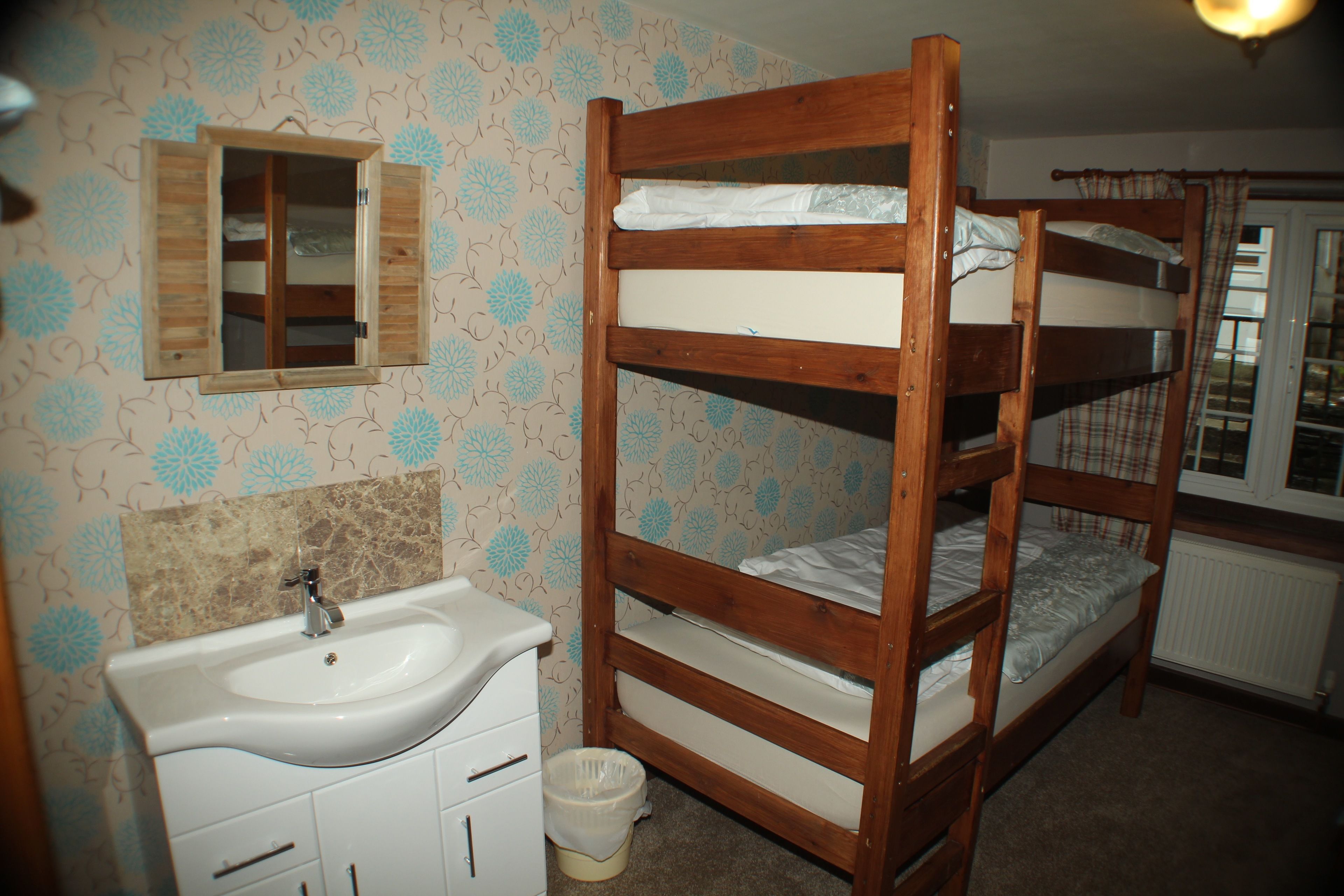 Un lit superposé avec une structure en bois et du linge de lit blanc, un lavabo blanc avec un miroir au-dessus, et un papier peint à motifs.