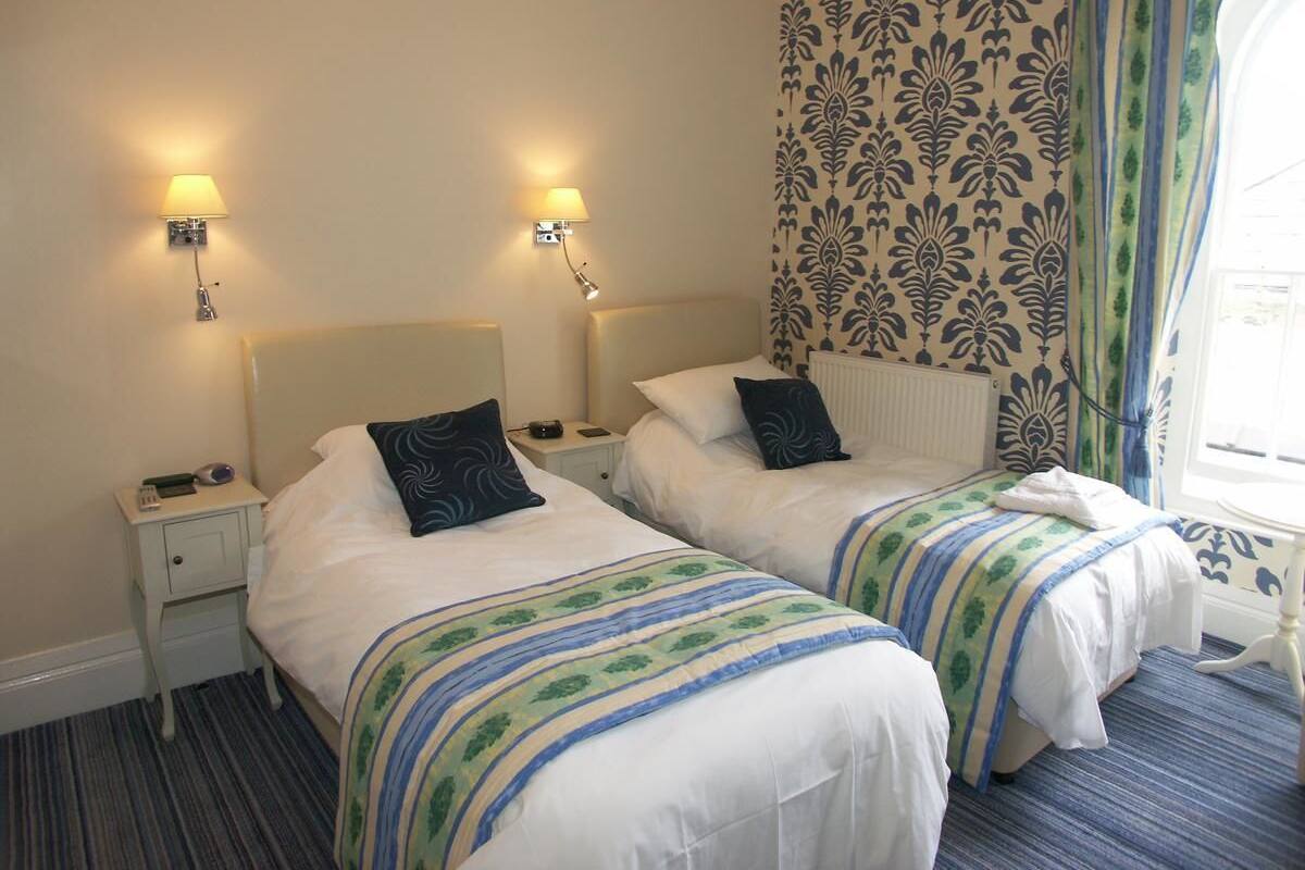 A hotel room with two beds, each with a headboard, bedside tables, lamps, and patterned curtains.