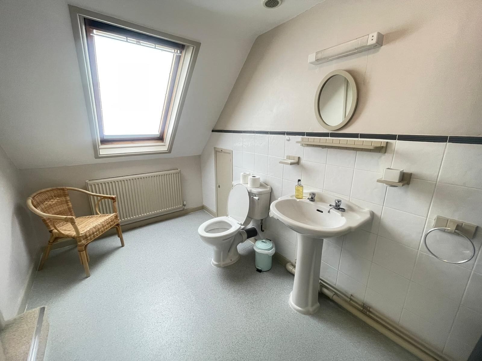 A small, modern bathroom with a toilet, bidet, and sink. There is a window providing natural light, a radiator, and a wicker chair.