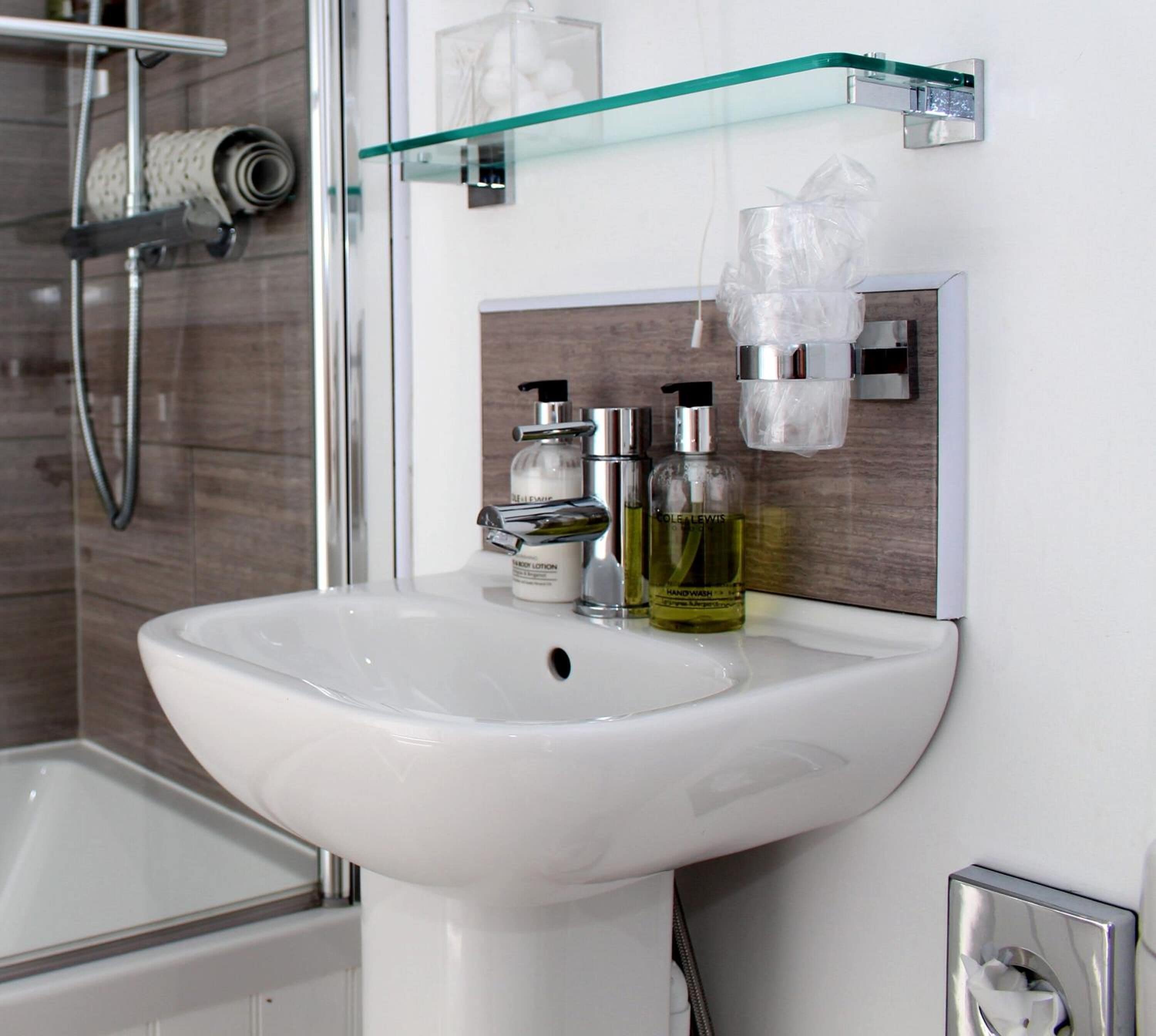 Une salle de bain avec un lavabo blanc, une étagère en verre où sont rangés les articles de toilette et un espace douche.