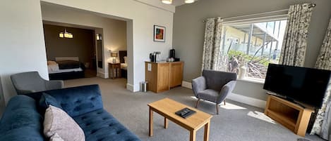 A modern living room with a large blue sofa, a wooden coffee table, a grey armchair, and a TV on a wooden stand. There is a small wooden cabinet and a lamp on a side table.