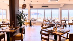 A spacious dining area with multiple tables and chairs, large windows offering a view of the sea, and a potted plant in the center. - The Cliff Hotel & Spa (Cardigan)