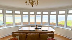 A room with a curved bay window overlooking a beach, a dining table, and a leather sofa with decorative pillows. - The Cliff Hotel & Spa (Cardigan)