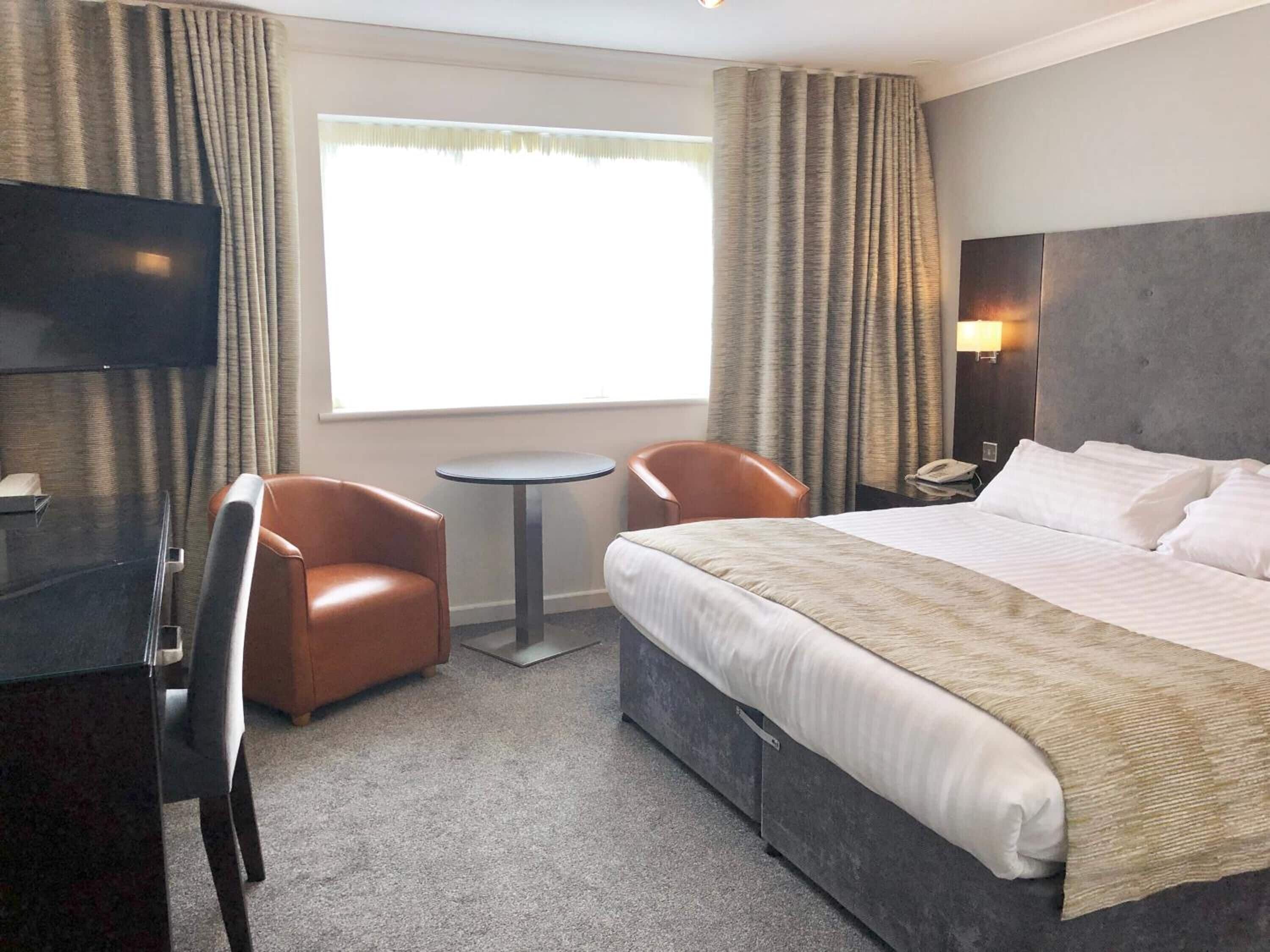 A hotel room with a large bed, two armchairs, a small table, a TV, and a window with curtains.