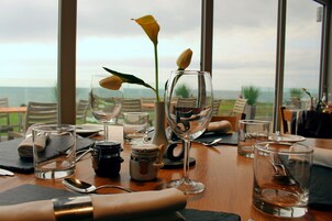 Una mesa lista para comer, con cristalería transparente, un florero con flores amarillas y vista al océano a través de amplias ventanas.