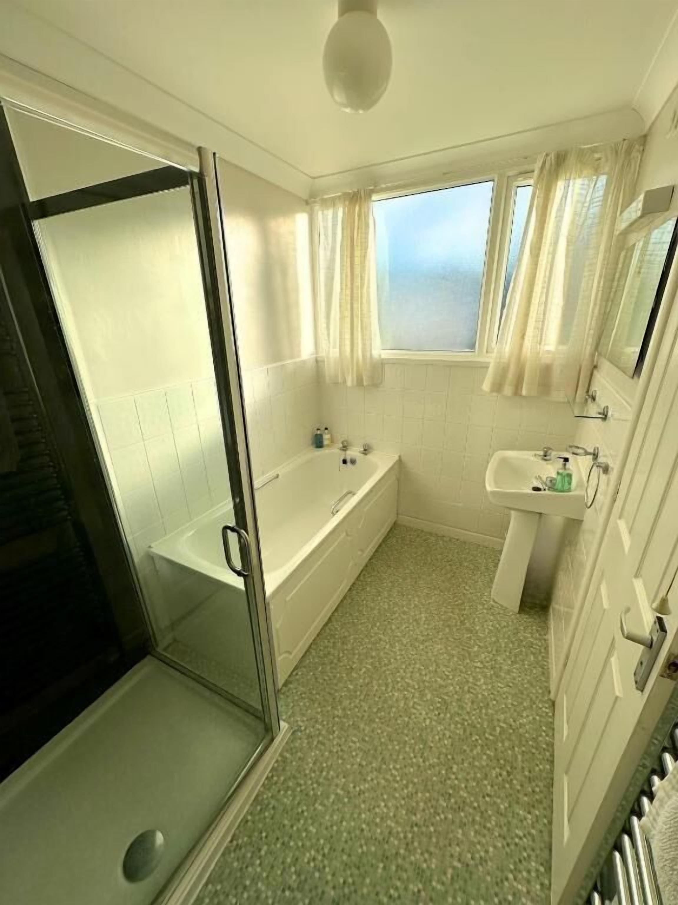 A bathroom with a glass shower enclosure, a bathtub, a sink, and a window with curtains.