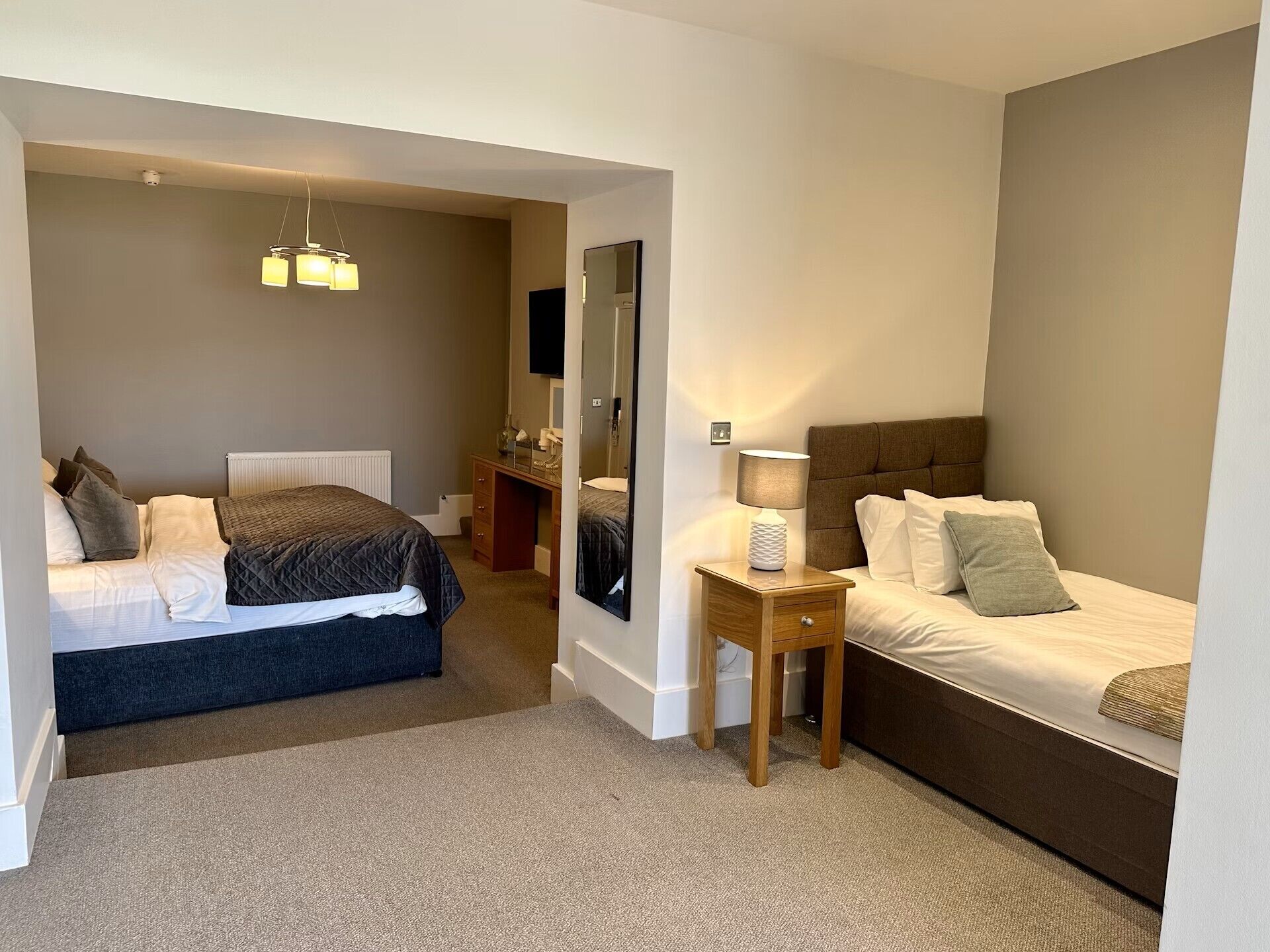Habitación de hotel con dos camas, una mesita de noche de madera con lámpara, un espejo y una lámpara en el techo.