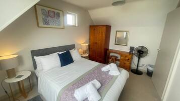 Ein Schlafzimmer mit Bett, Nachttisch, Lampe, Kleiderschrank, Schreibtisch, Stuhl und einem Standventilator.