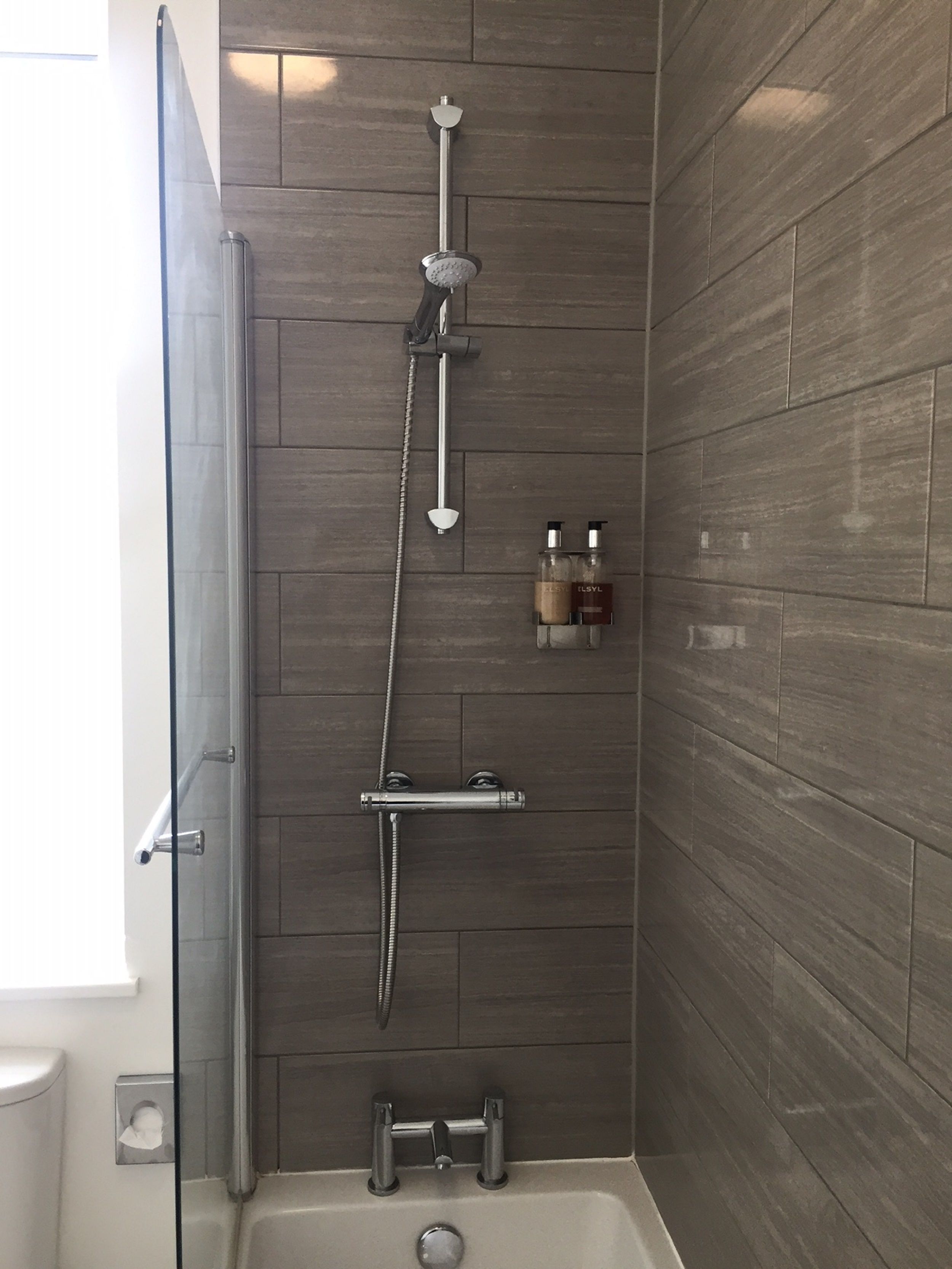 Ducha con cabezal de ducha manual, grifo empotrado en la pared y un cabezal de ducha manual. Hay un estante pequeño con dos botellas.
