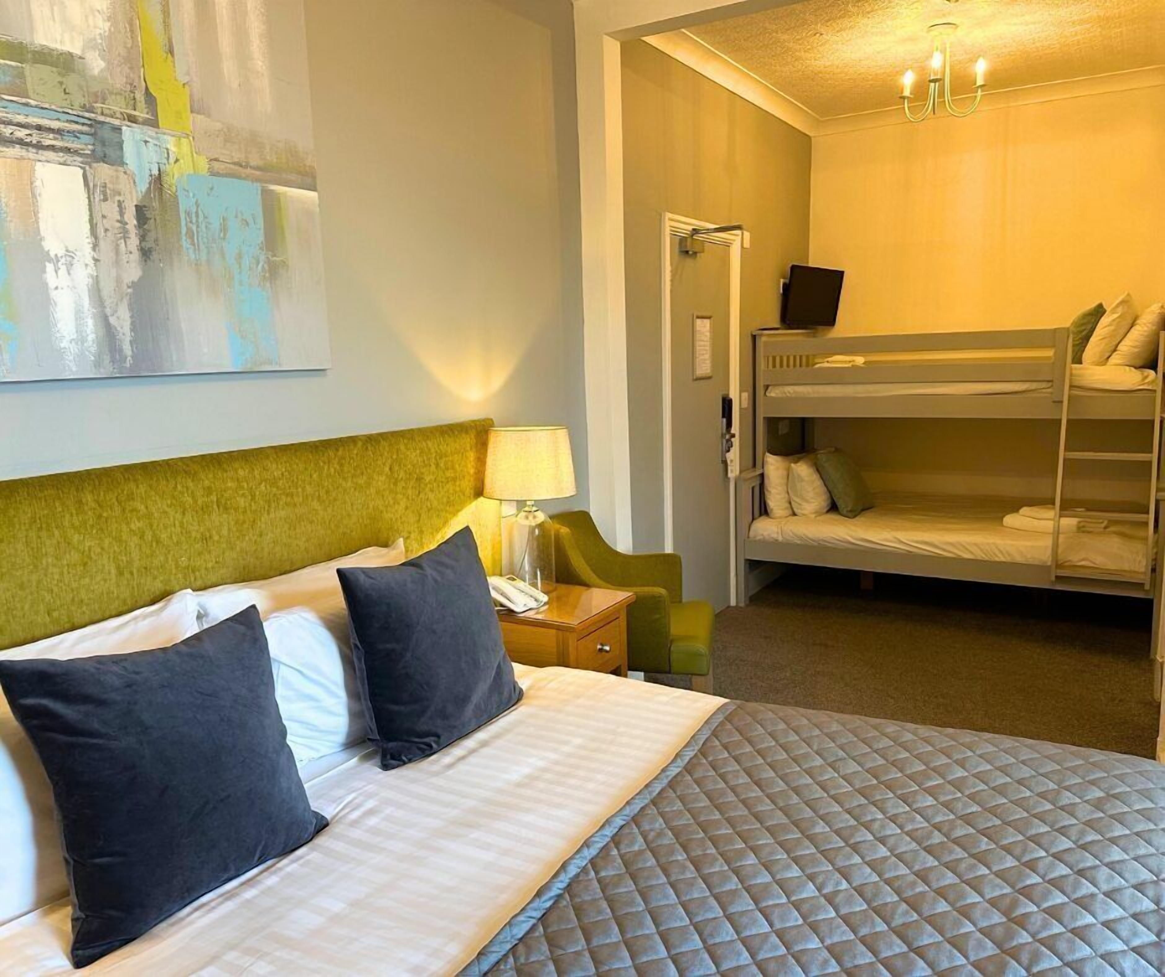A hotel room with a single bed, a bunk bed, a bedside table with a lamp, and a painting on the wall.