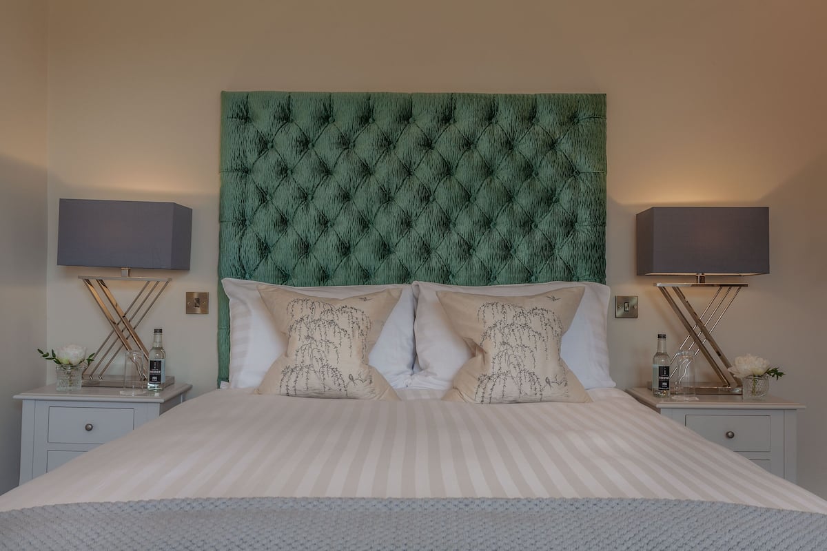 A neatly made bed with striped bedding, a tufted green headboard, and two bedside tables with lamps and small vases.
