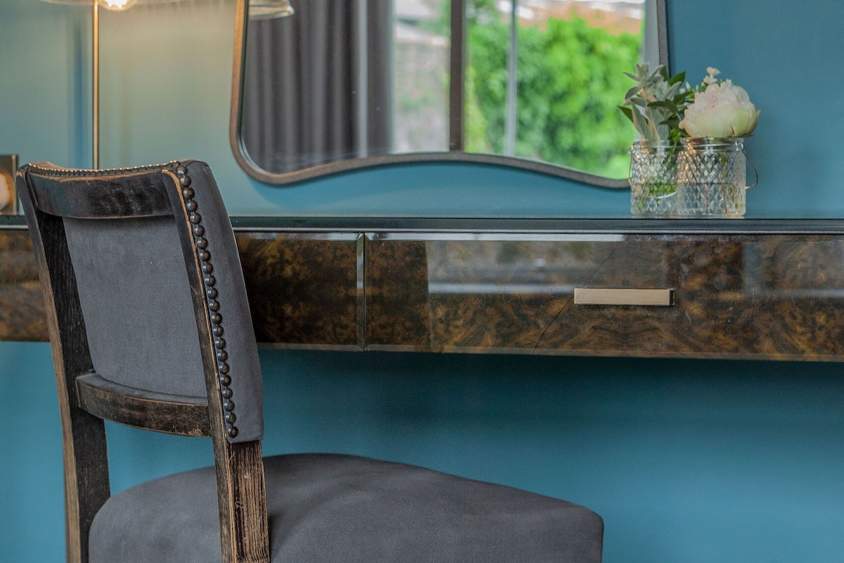 A chair with a dark wooden frame and a grey upholstered seat, positioned in front of a blue wall with a mirror reflecting a window view.