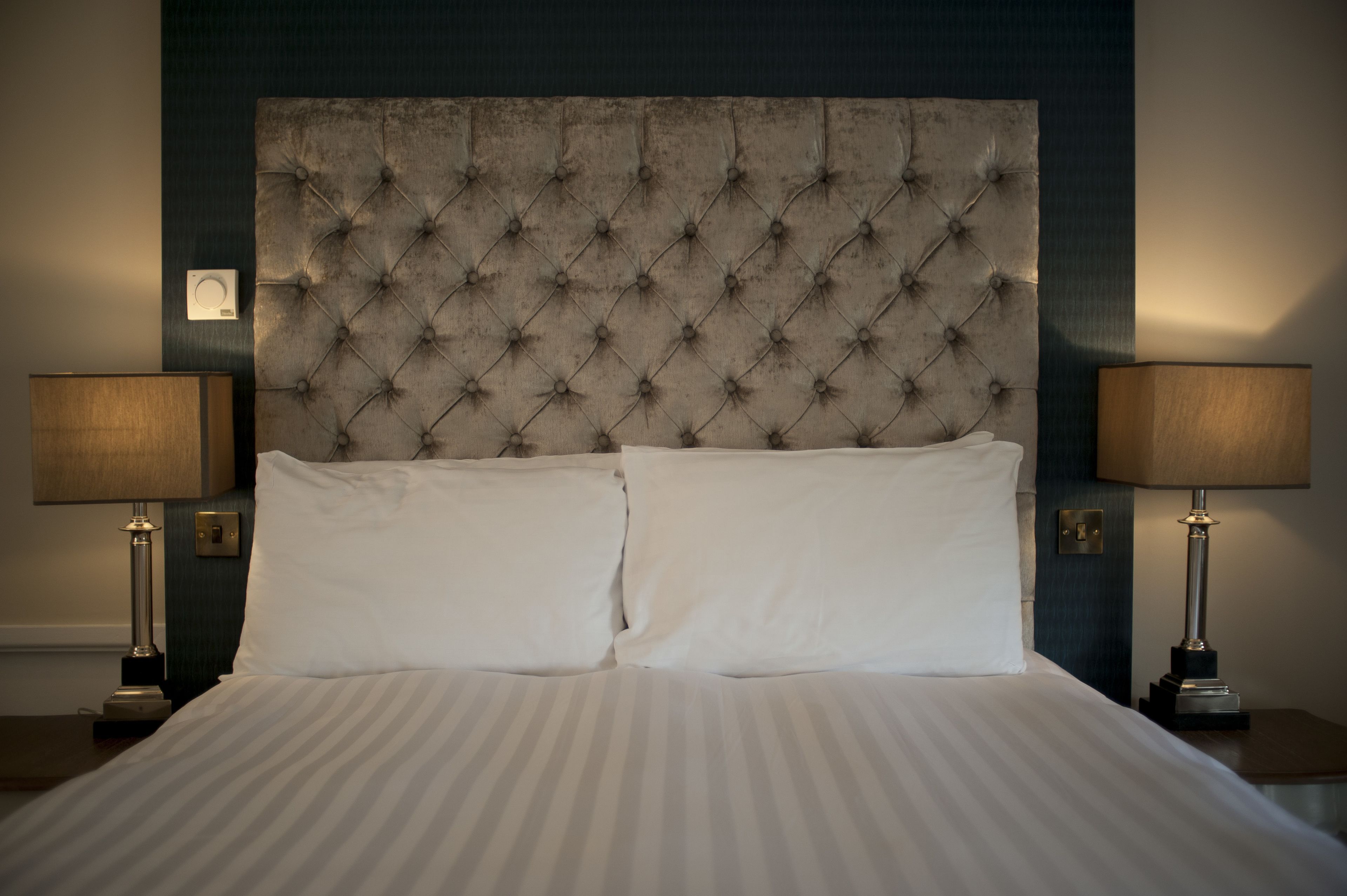 Quarto de hotel com cabeceira estofada, duas luminárias de apoio e cama bem arrumada com lençóis brancos.