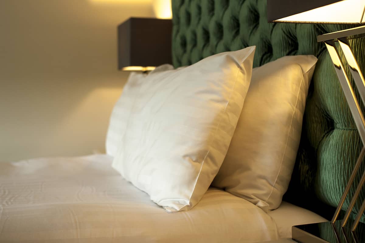 A neatly made bed with white linens and two pillows, a green upholstered headboard, a bedside table with a lamp, and a nightstand with a drawer.